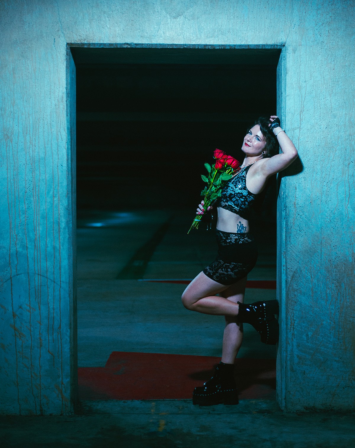 Person holding red flowers, standing in doorway, posing with one leg raised, wearing black clothing, dark background.