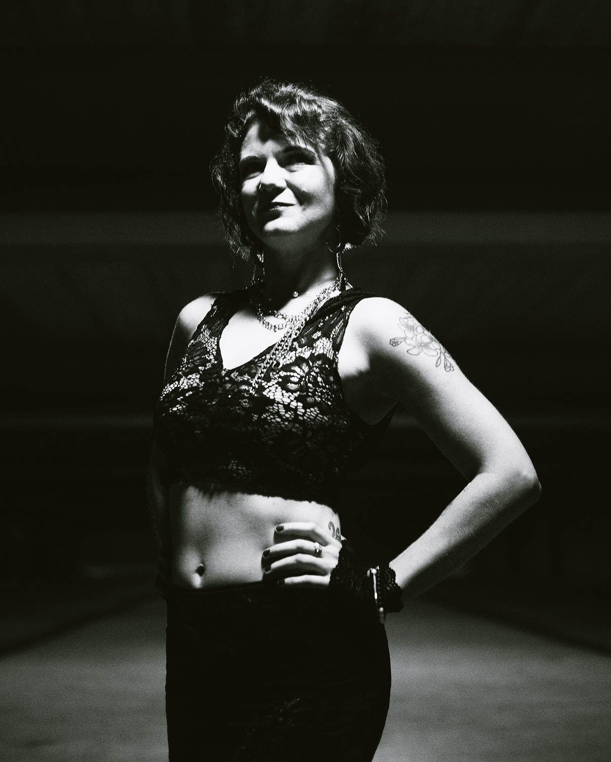 Woman with short curly hair wearing a lace crop top, standing with one hand on her hip, smiling, in a dark setting.