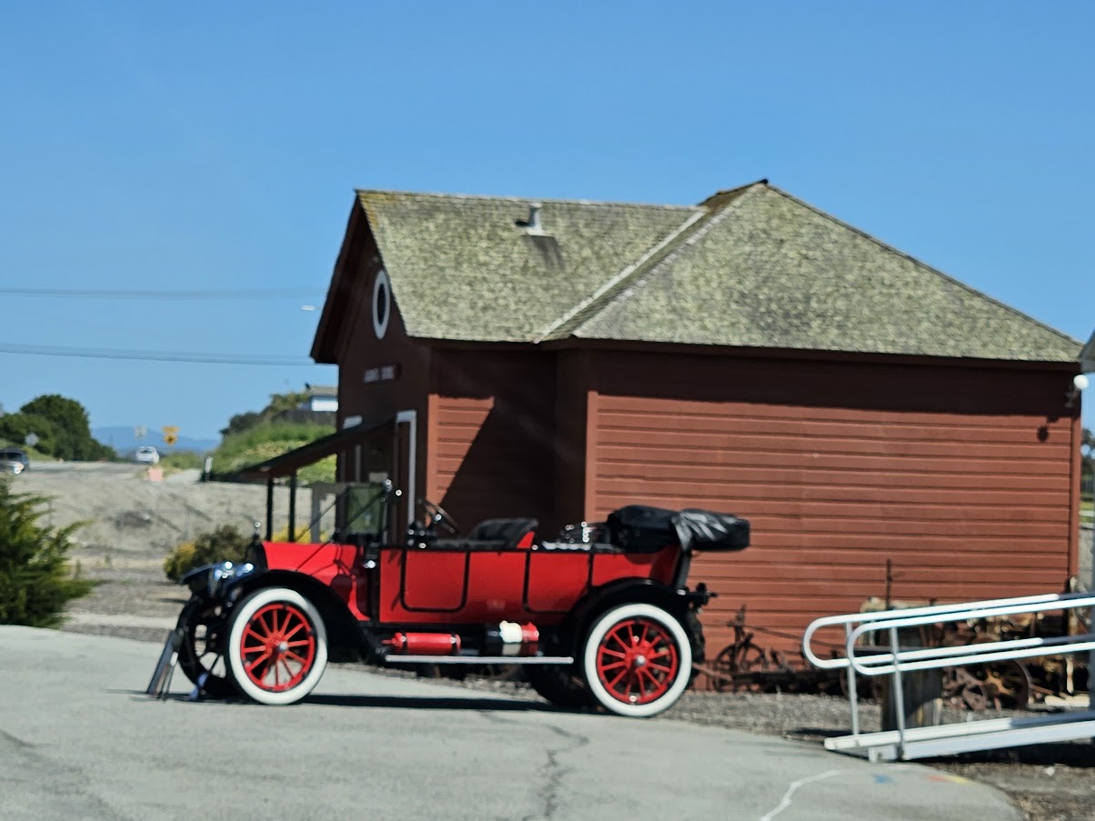 Red vintage car parked in front of a red wooden building with a sloped roof, under a clear blue sky.