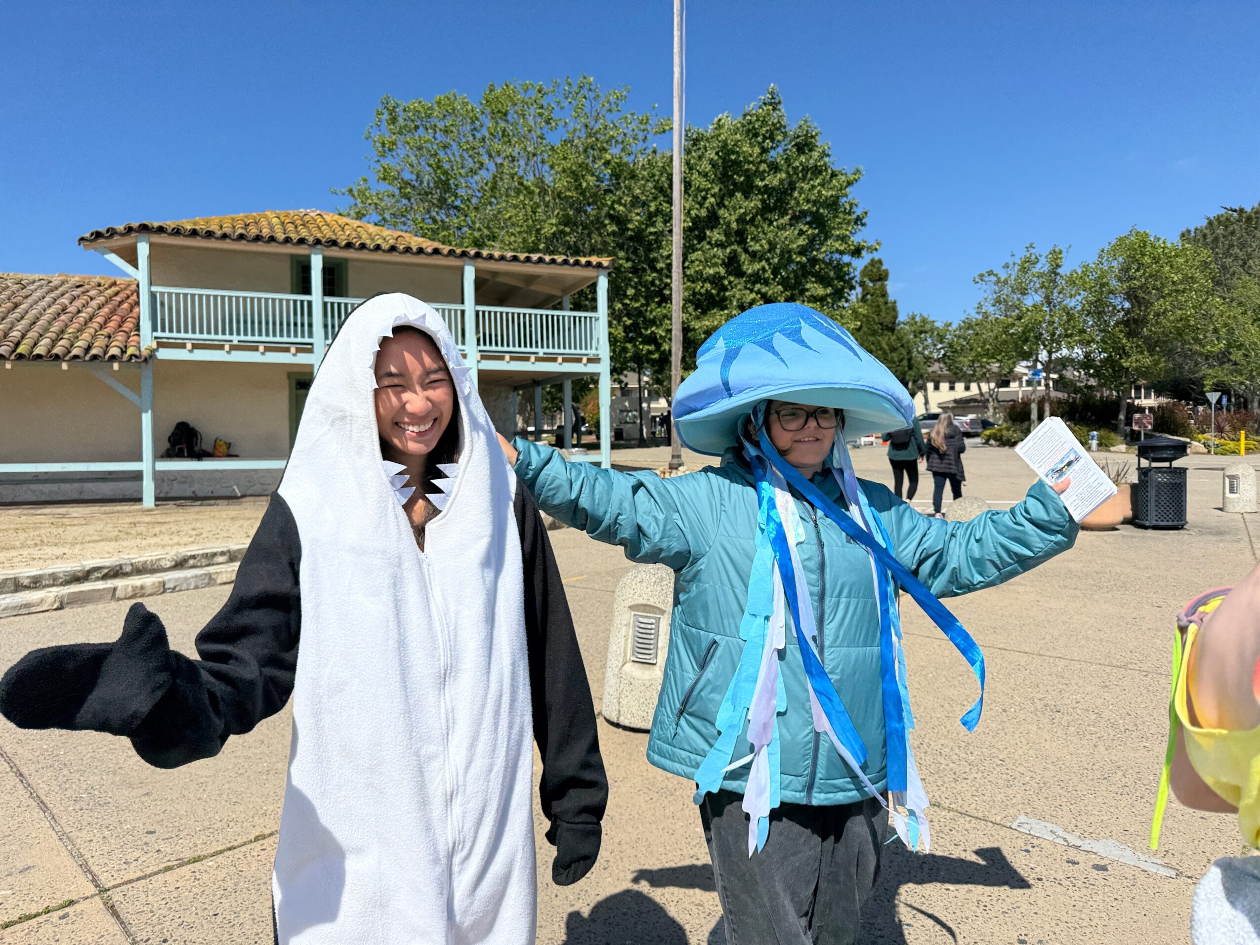 Two children wearing large blue hats and jackets, smiling outdoors with a building and trees in the background.