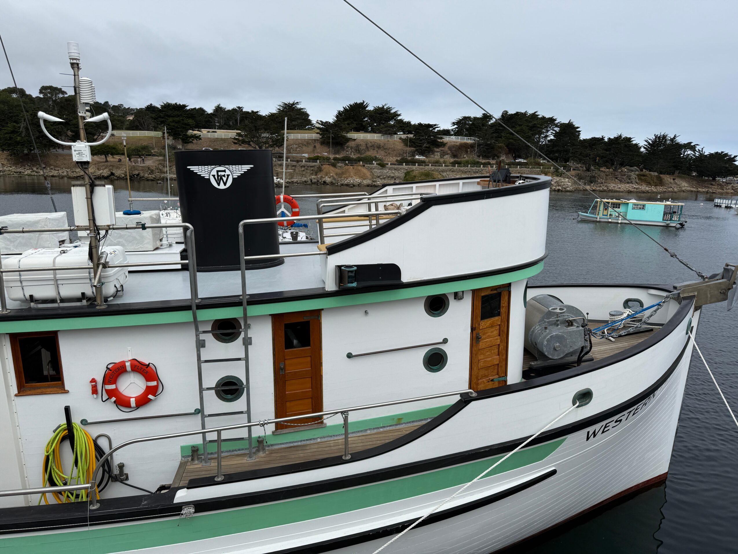 Boat docked at a marina with water and other boats in the background, featuring a black water tank, life ring, and wooden doors.