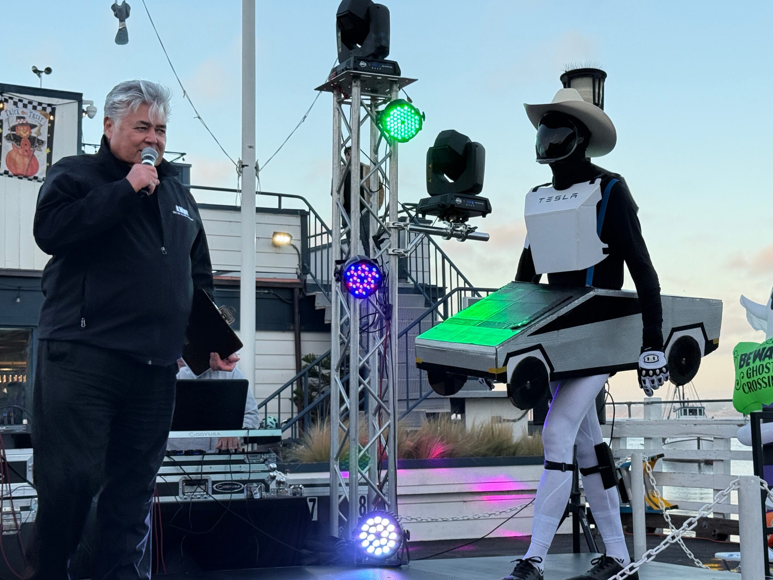 Man speaking into microphone on stage with a person in a robot costume holding a car model, stage lights, and boat in background.