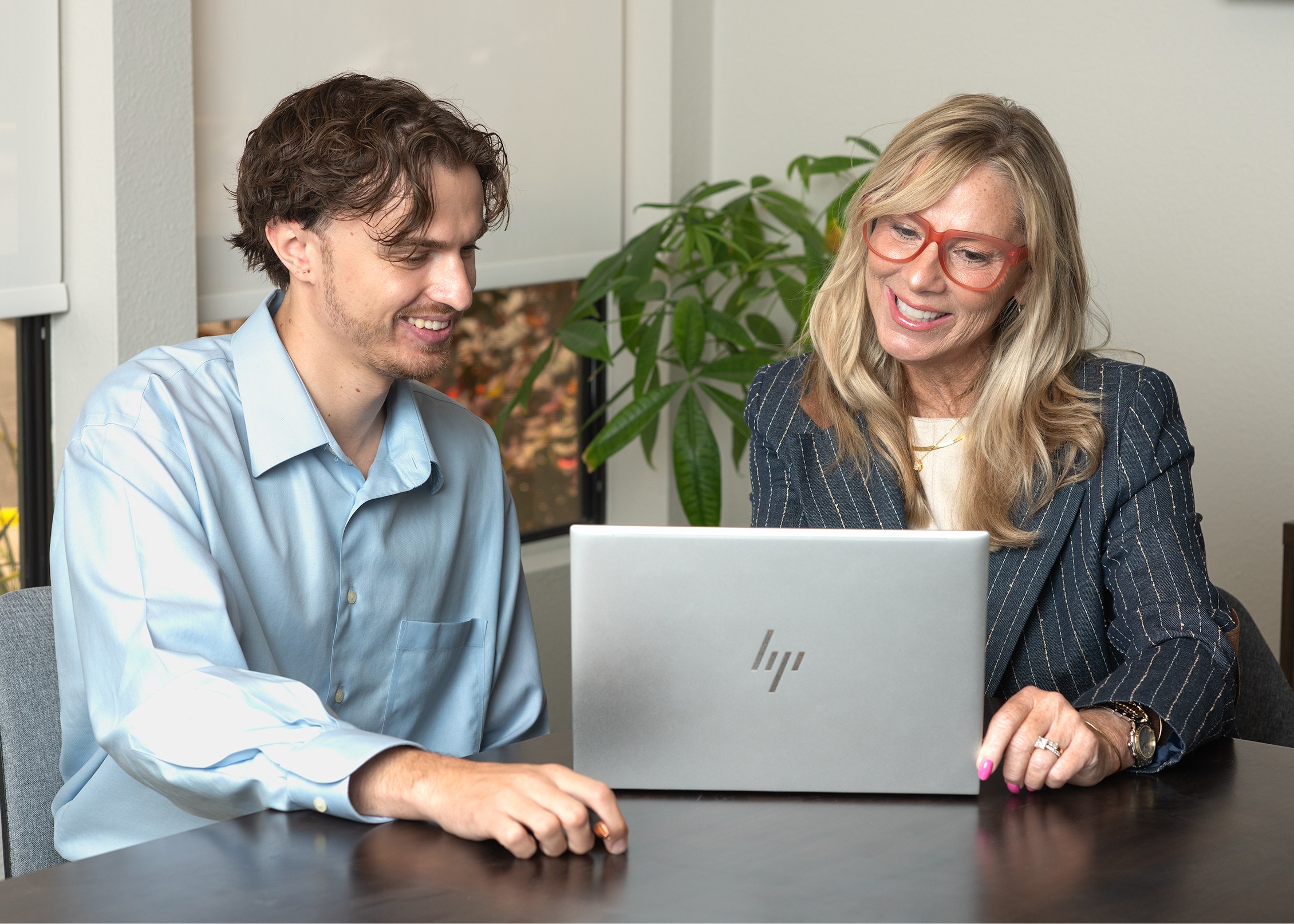 Two people sitting at a table looking at a laptop, smiling, with a plant in the background.