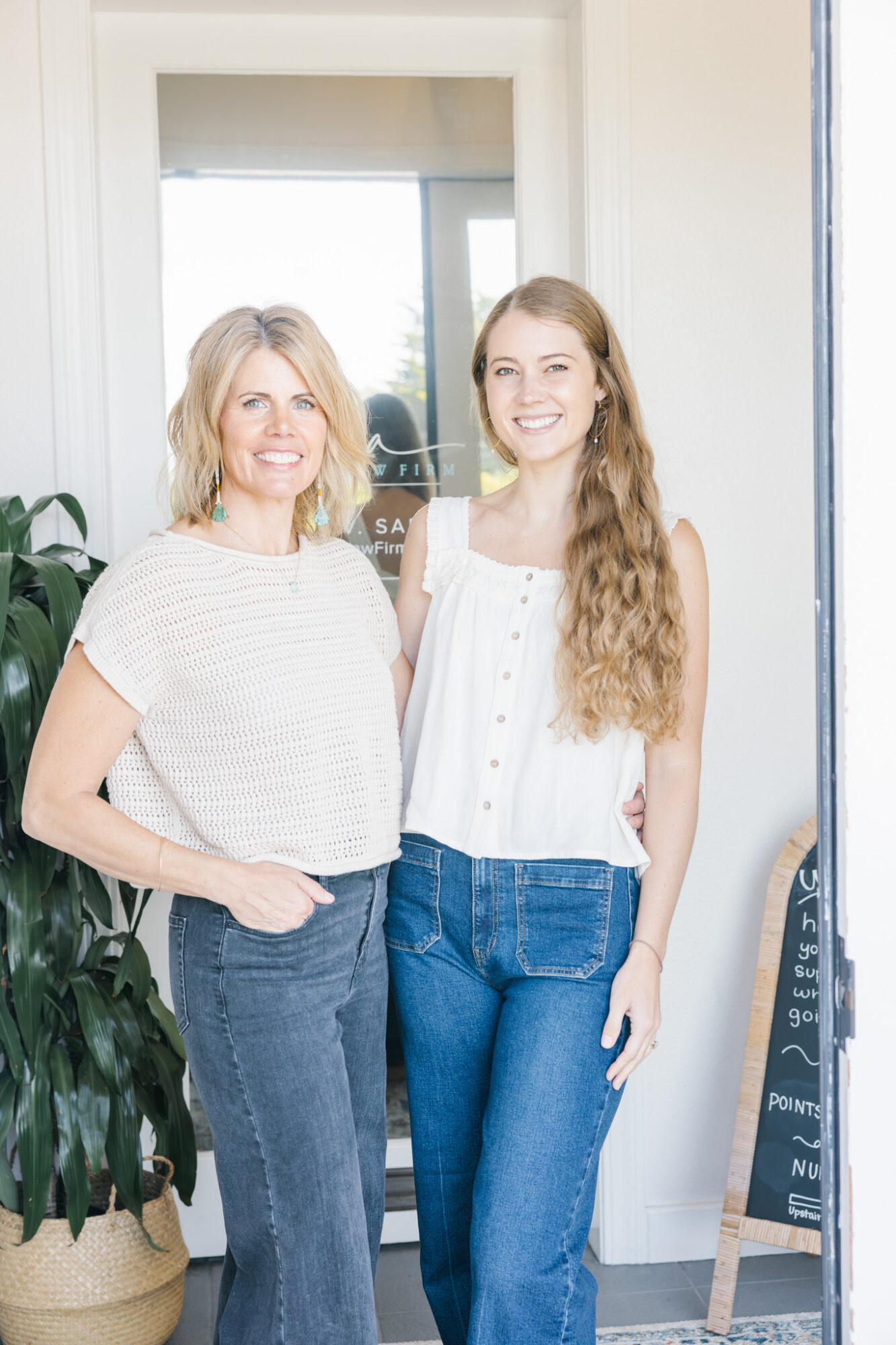 Two women standing indoors, smiling, with a window and a chalkboard sign in the background.