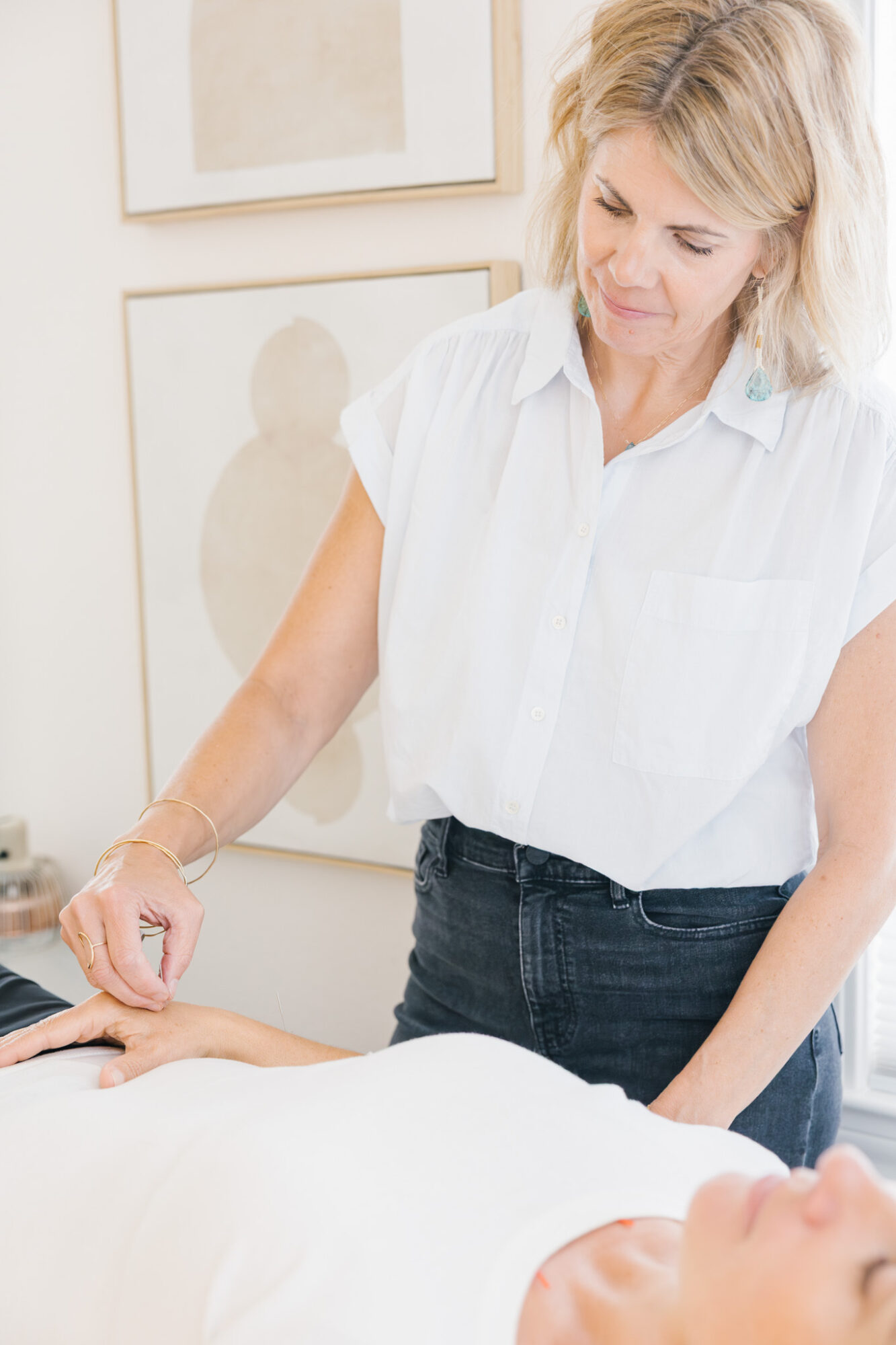 Woman with blonde hair in a white shirt leaning over a person lying down, in a room with framed artwork on the wall.