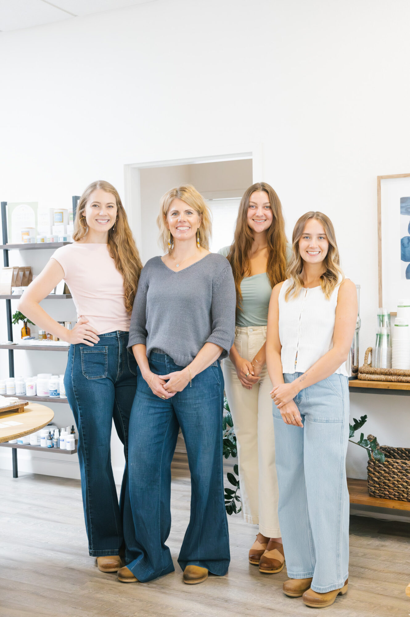 Four women standing together in a bright room, smiling, with shelves and framed pictures in the background.