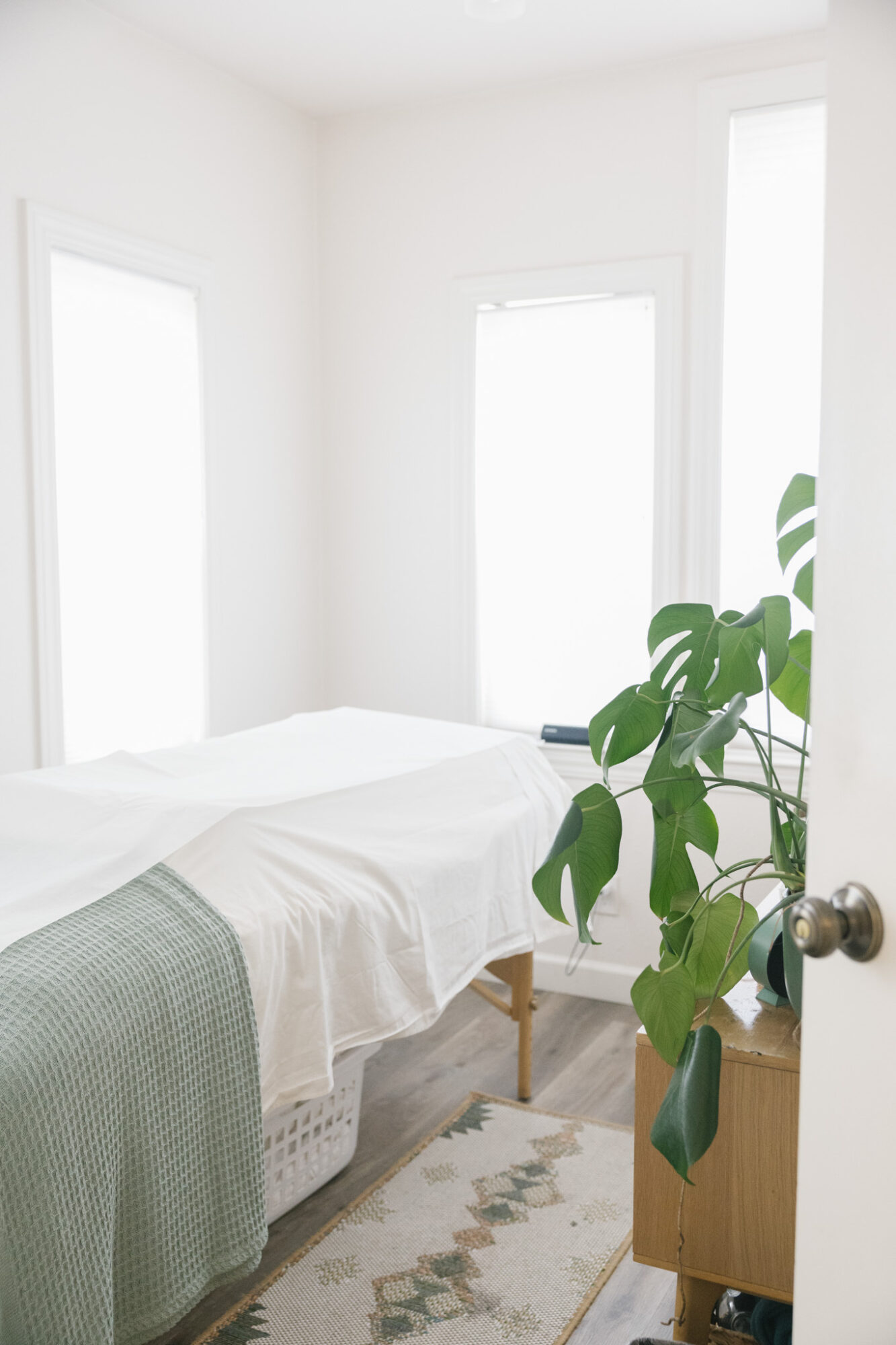 Bright room with white walls, large windows, a bed with white bedding, a green blanket, and a wooden cabinet with a plant.