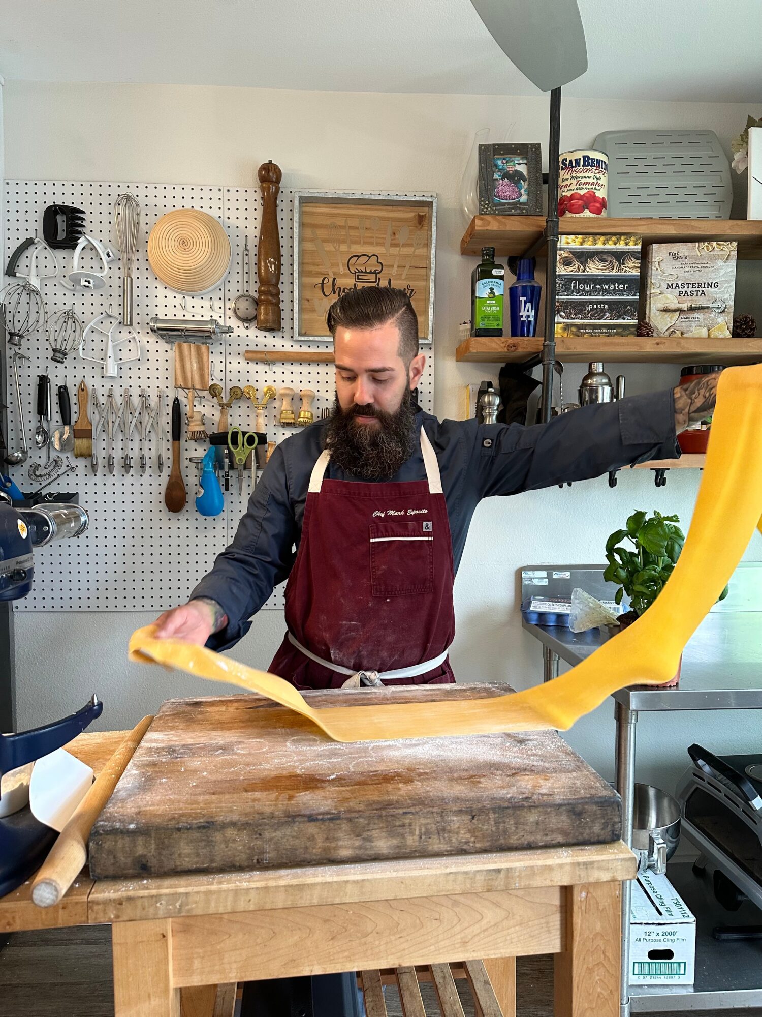 Man with beard in apron stretching yellow dough on a wooden table in a bakery or kitchen.