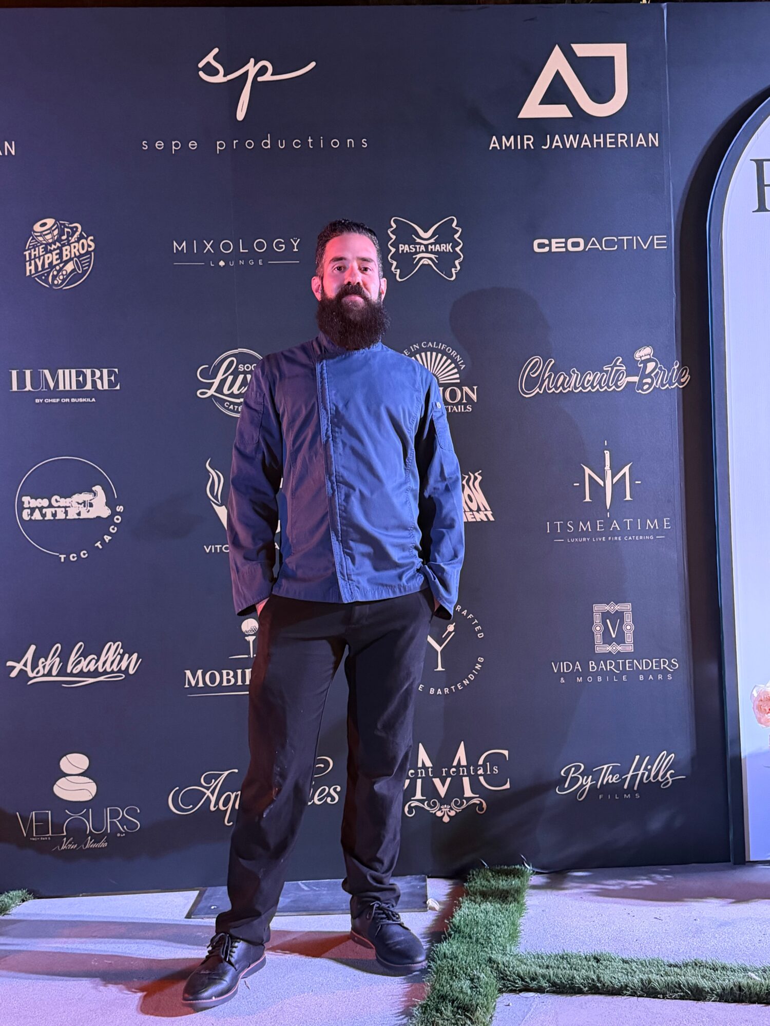 Man with beard in blue and black jacket standing on stage with a dark backdrop featuring various logos.
