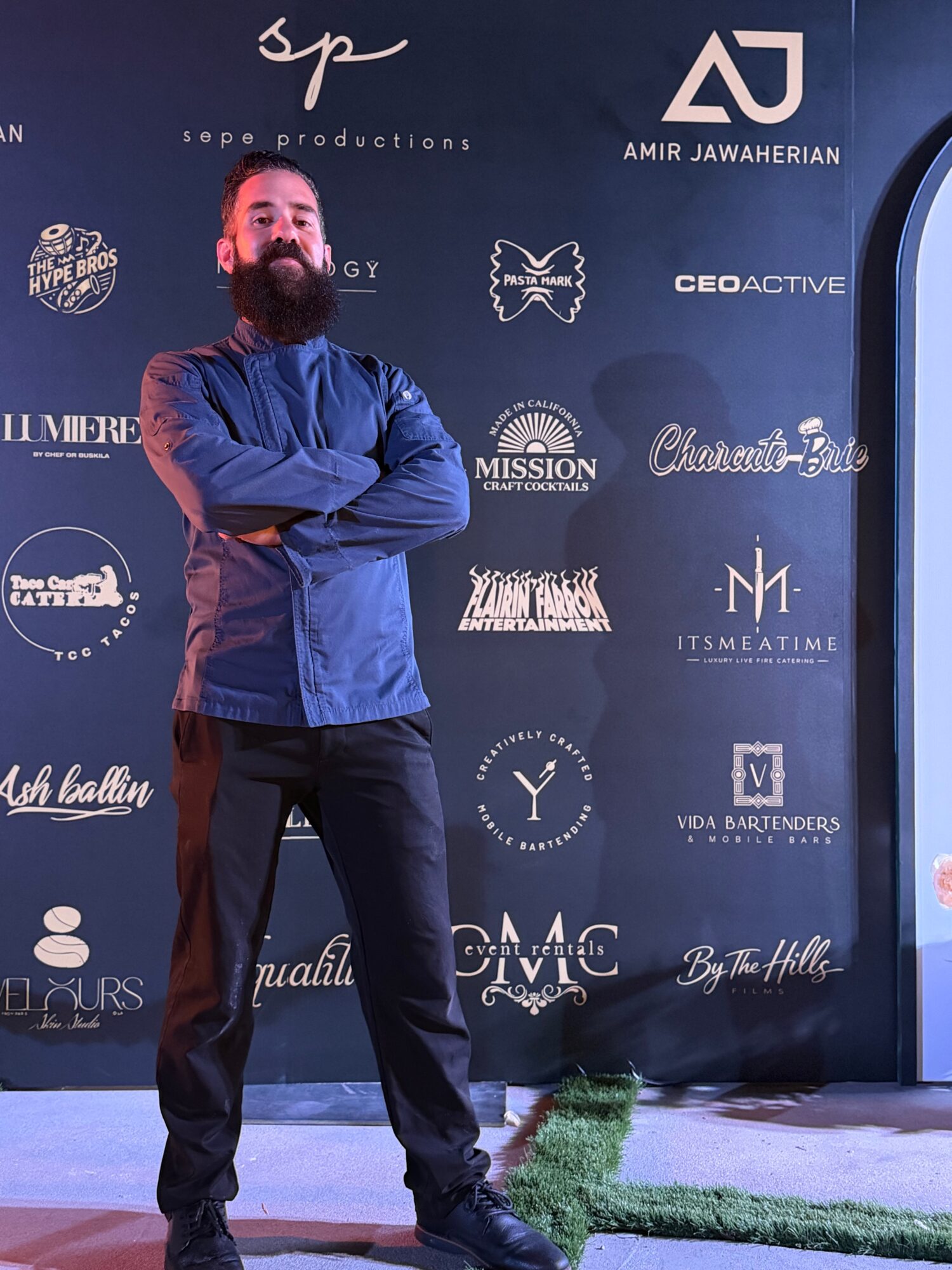 Man with beard and crossed arms standing in front of a dark backdrop with logos and text.