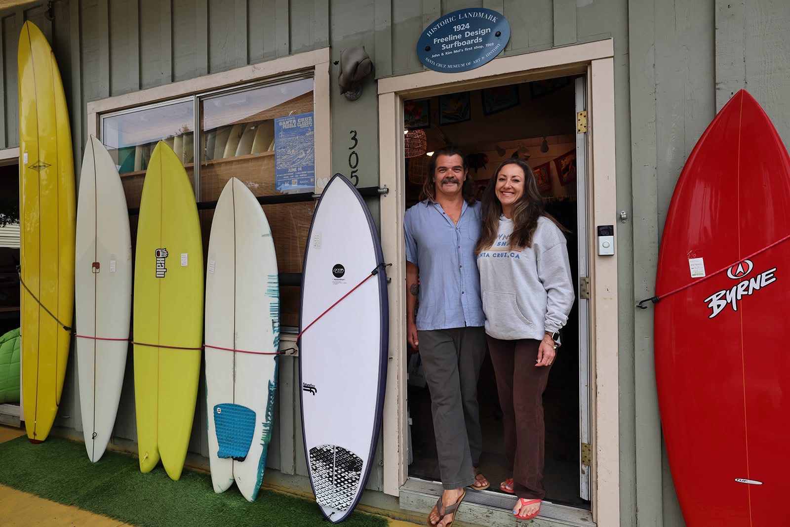 Two people stand in doorway of surf shop with surfboards outside, smiling. Shop has a blue sign above door.