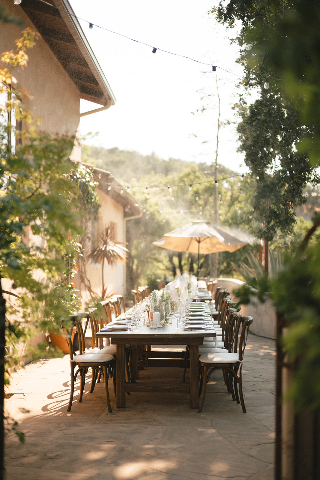 Outdoor dining table with chairs, umbrella, and string lights, surrounded by trees and buildings, sunny day.