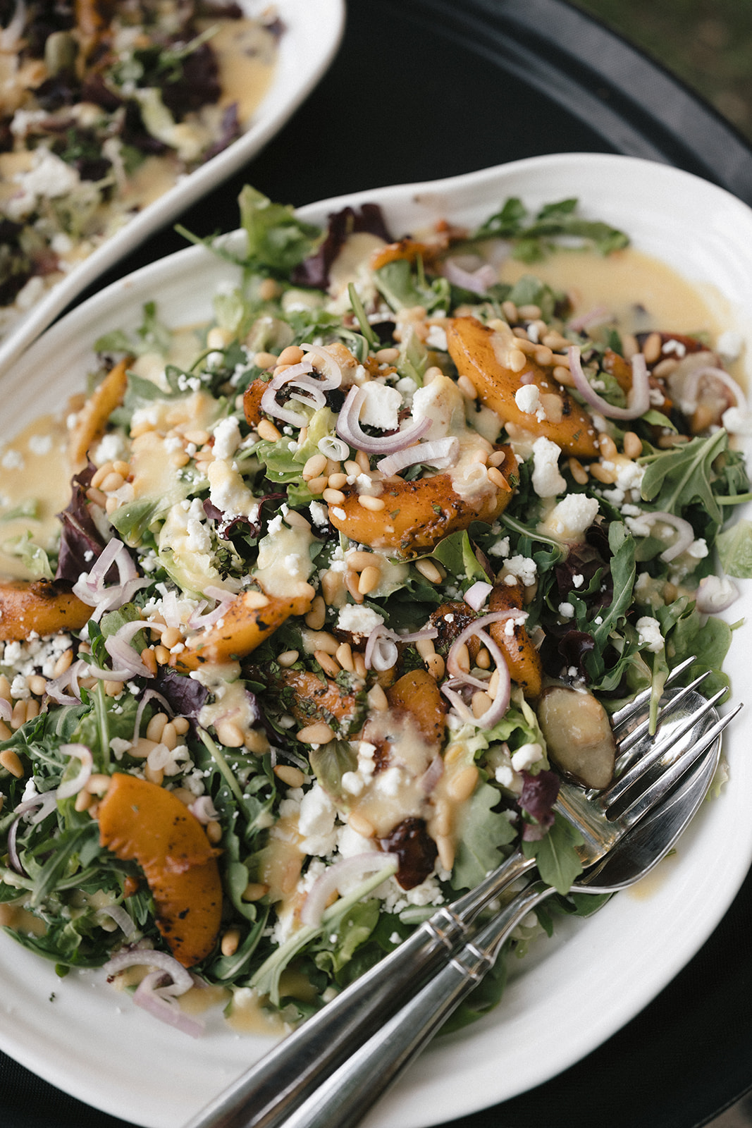 Salad with greens, roasted vegetables, cheese, and chopped onions on a white oval plate, with a fork and spoon, close-up.
