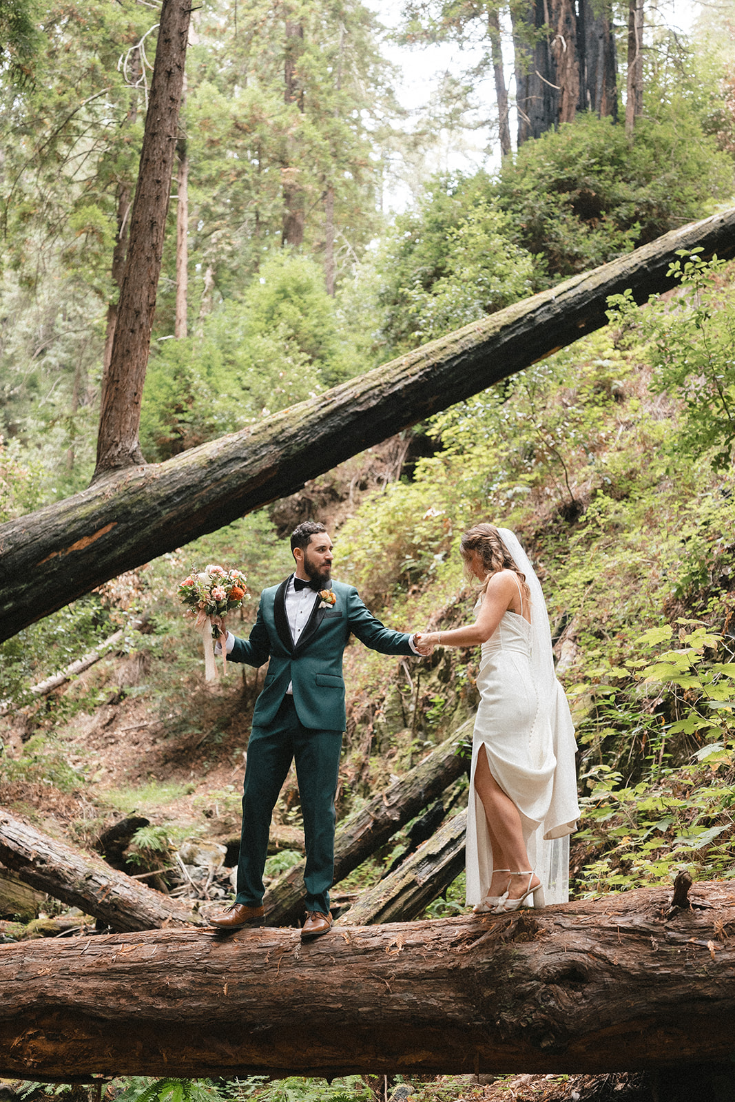A couple in wedding attire holding hands in a forest with fallen trees and green foliage.