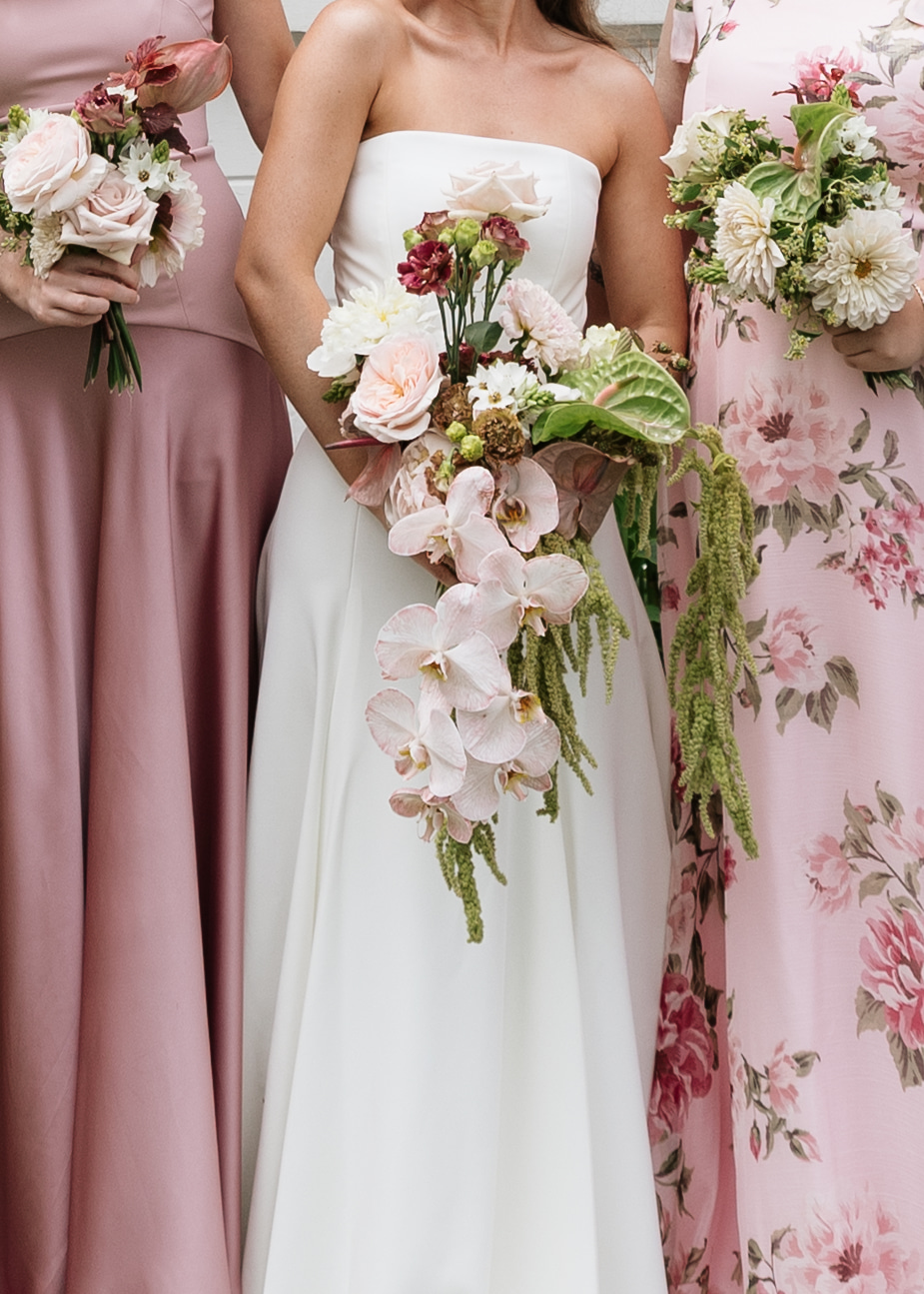 Three women in floral and solid-colored dresses holding bouquets, with the central woman in a white strapless dress, holding a large cascading bouquet.