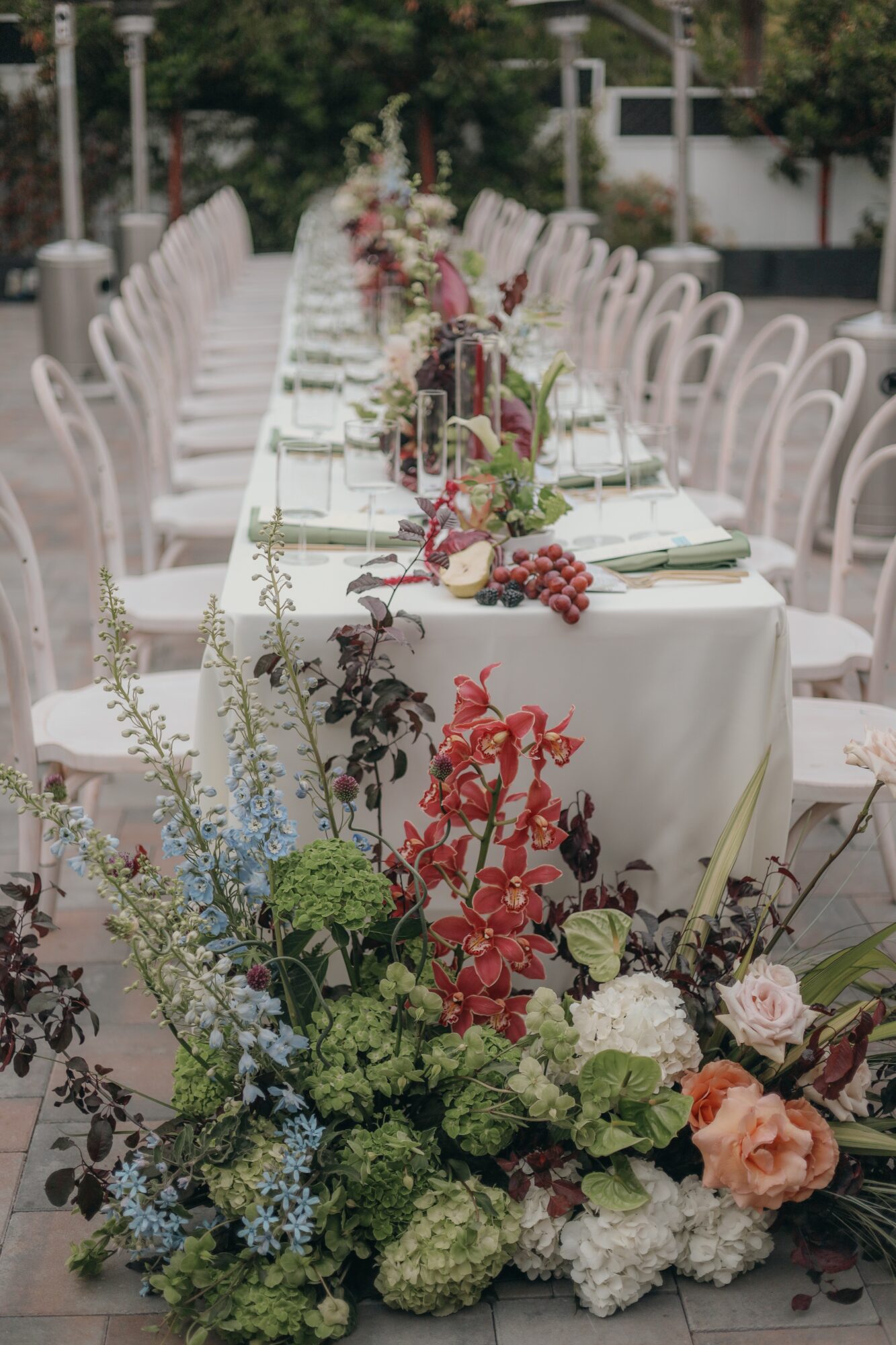 Long outdoor table decorated with floral arrangements, white chairs, and table settings, set for an event or celebration.