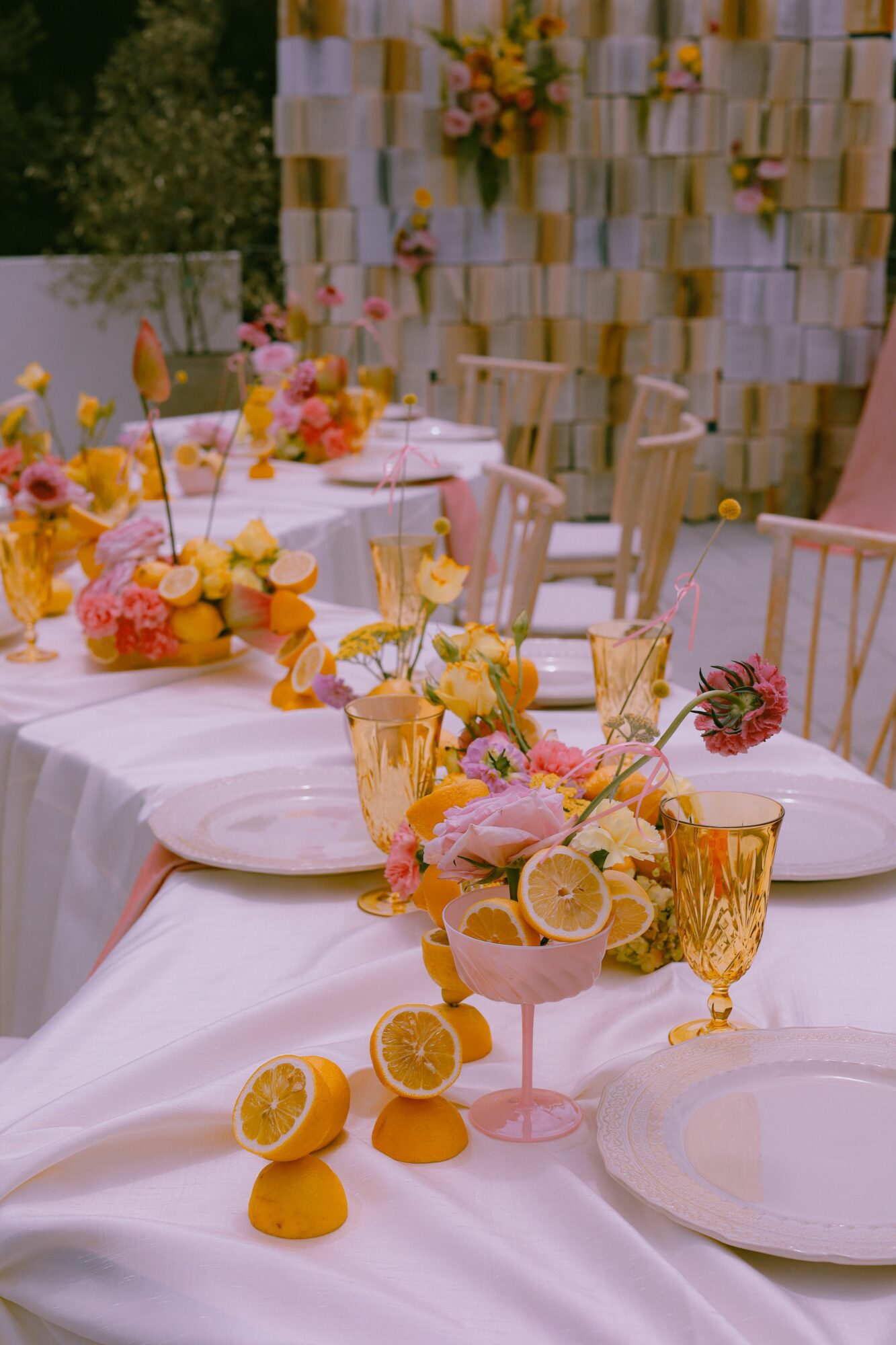 Decorated table with flowers, lemons, and gold-rimmed glasses at an outdoor event, with a floral backdrop.