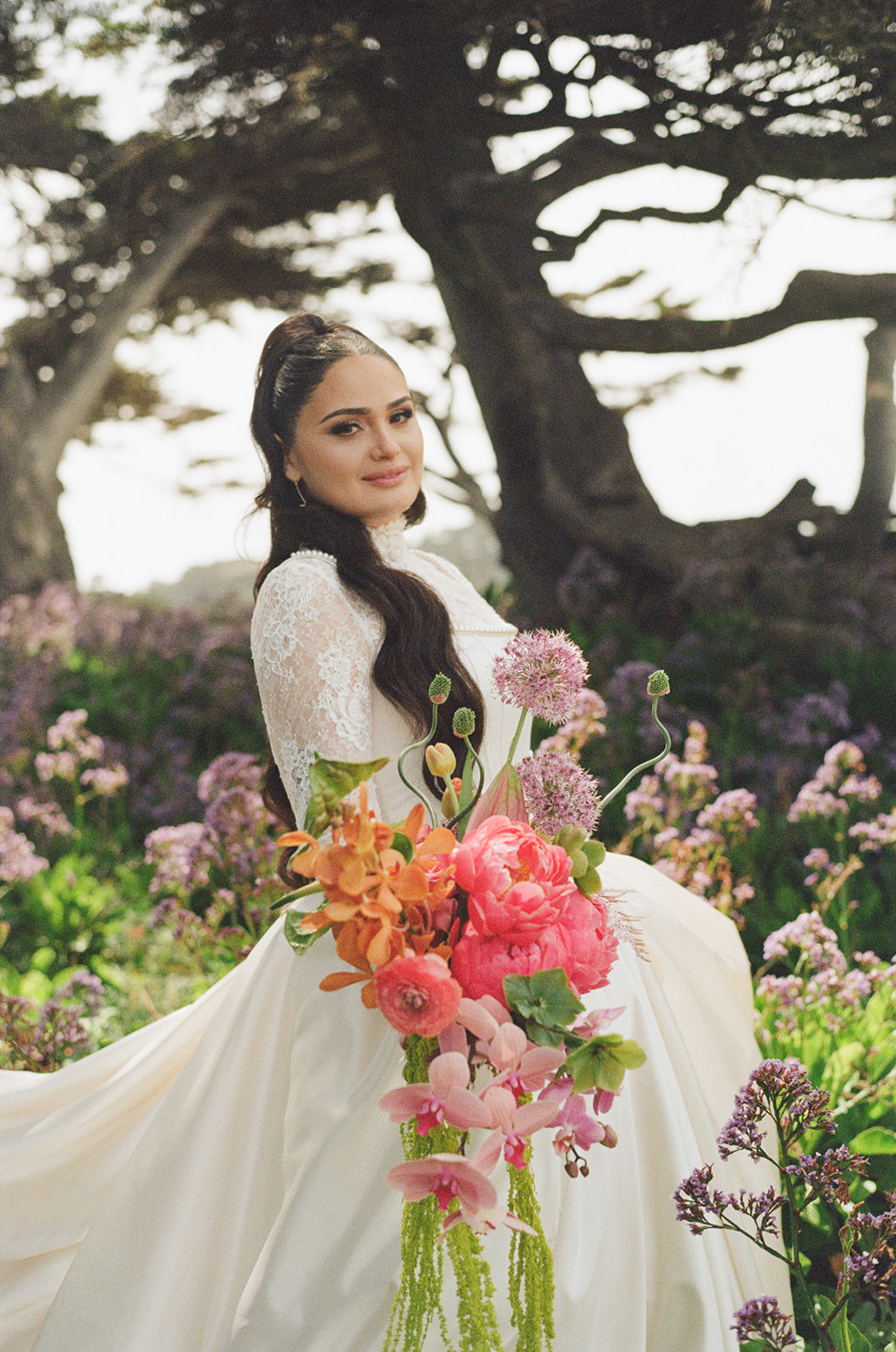 Woman in a white dress holding a large bouquet of pink and orange flowers outdoors, with trees and flowers in the background.