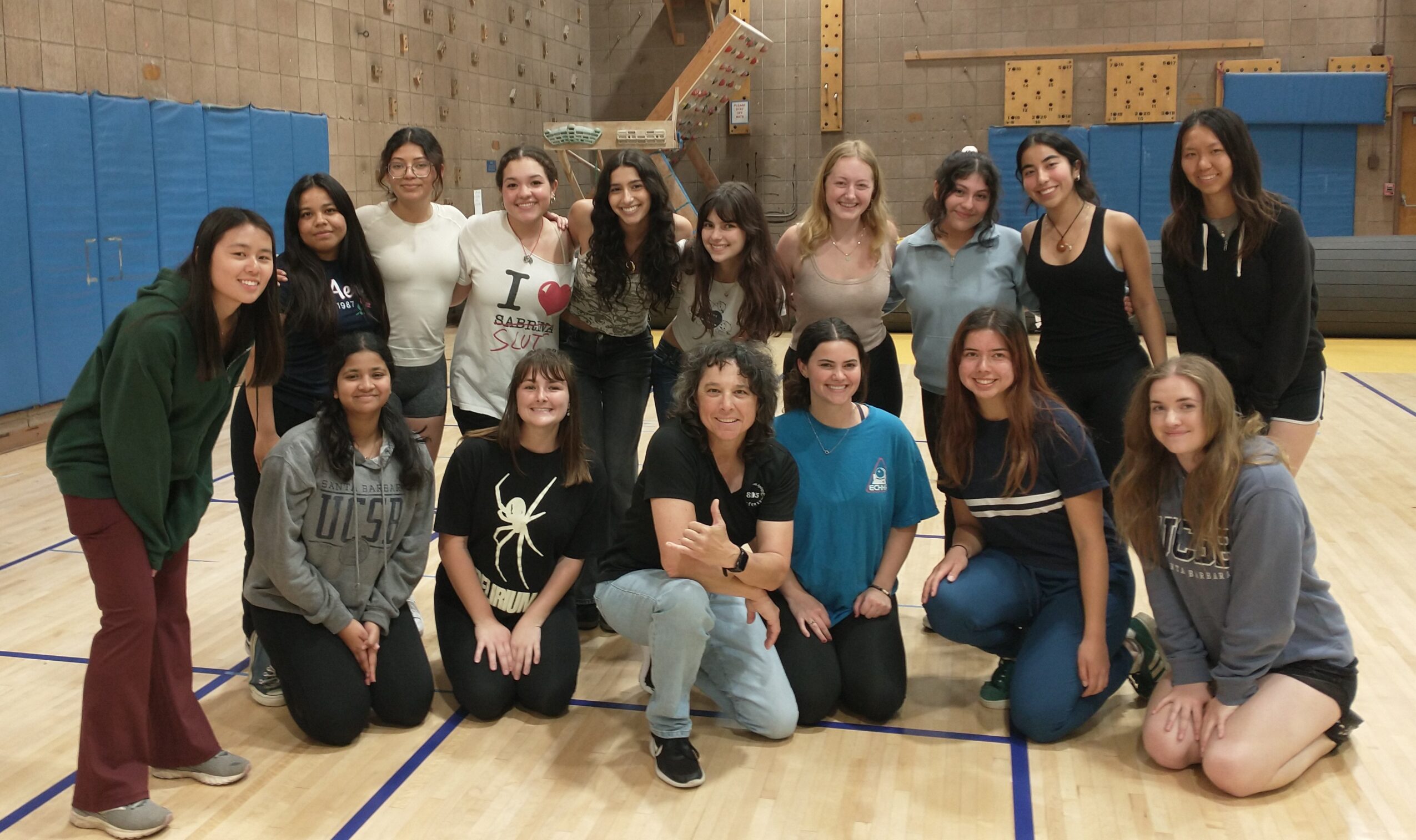 Group of 16 young women posing in a gymnasium, some standing and some kneeling, smiling at the camera.