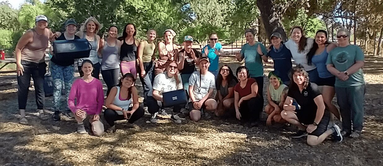 Group of people outdoors, some standing and some sitting, with trees and a grill in the background.