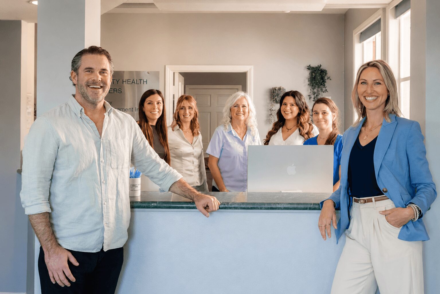 Group of six people smiling in a bright office, with two in the foreground and four in the background, behind a reception desk.