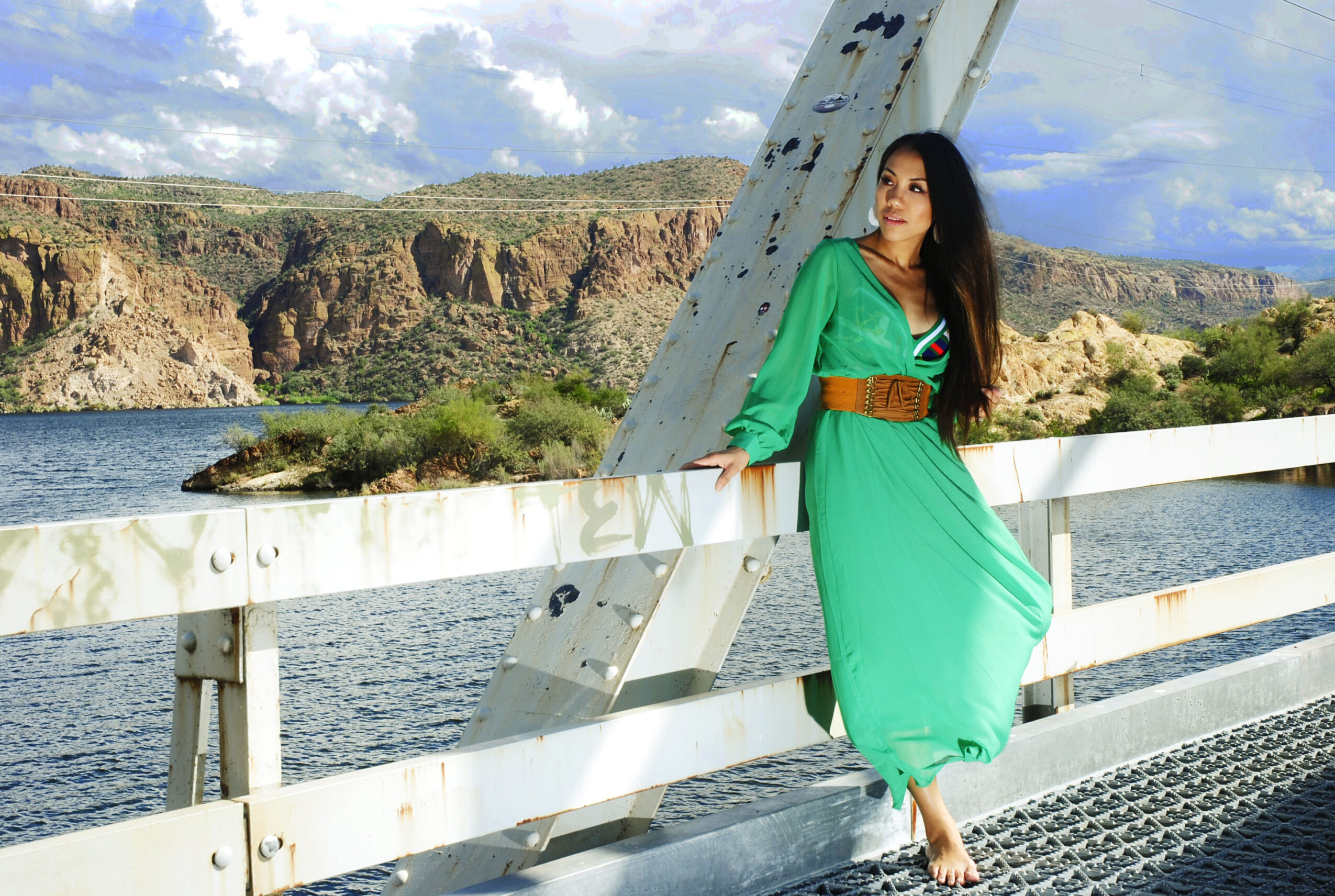 Woman in green dress standing on bridge with river and rocky hills in background.