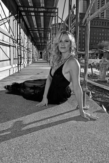 Woman sitting on sidewalk with construction scaffolding and city buildings in background, sunlight casting shadows.