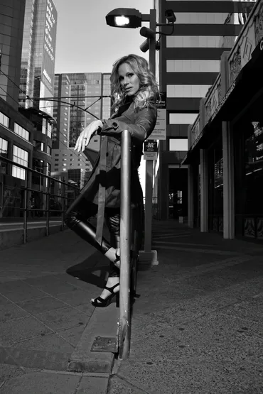 Woman leaning on a pole on city street at night, buildings and streetlights in background.