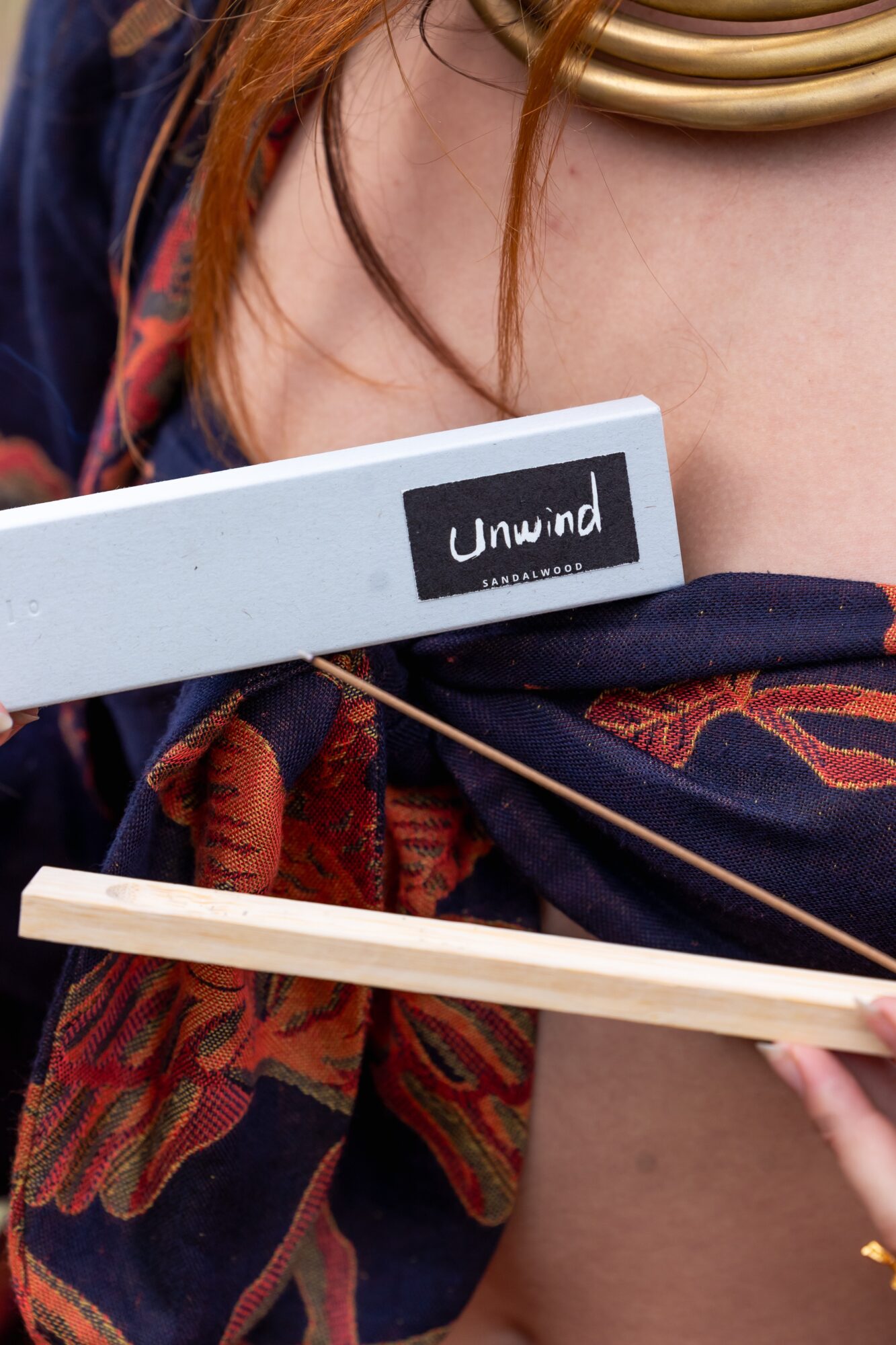 Close-up of a person holding a white box labeled 'Unwind' and a wooden stick, with a patterned scarf and gold necklace visible.