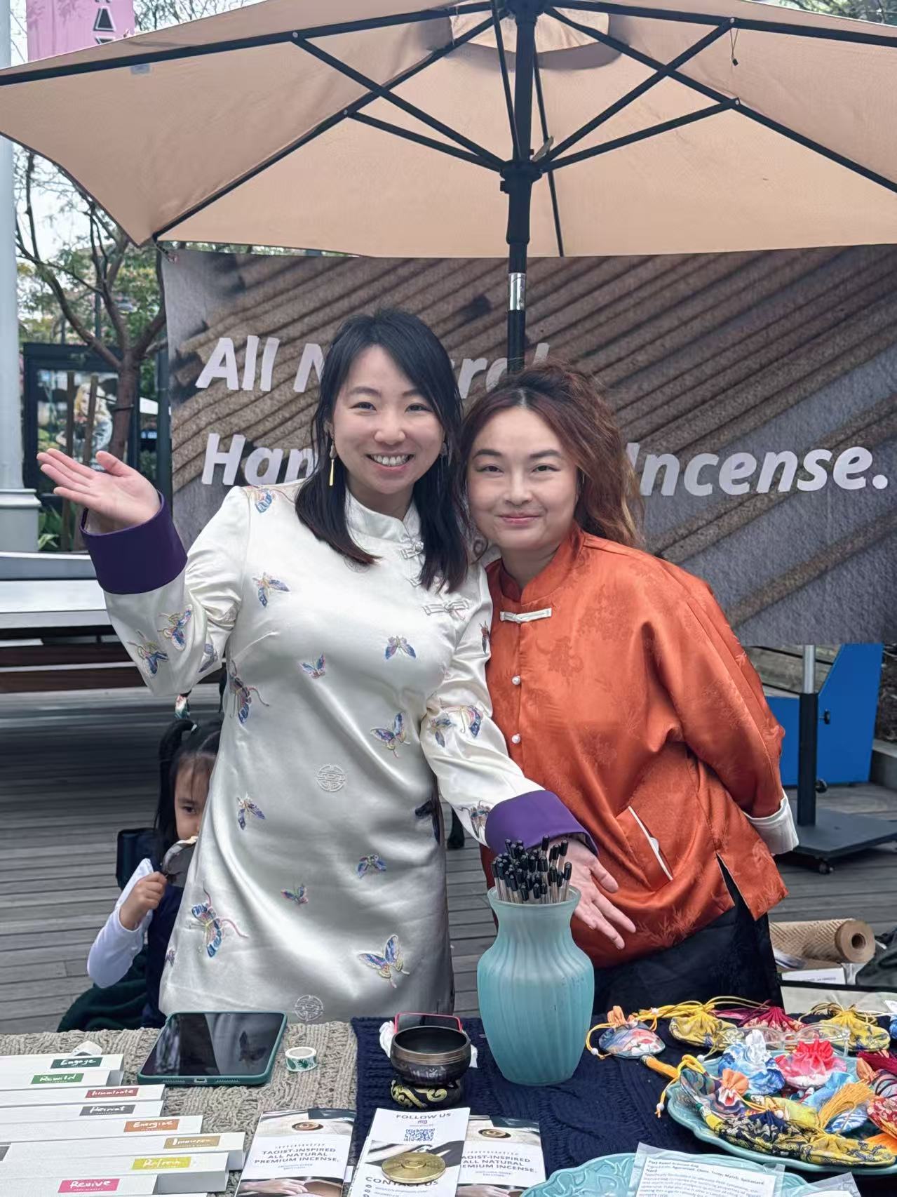 Two women smiling and posing outdoors under a large umbrella, with a table of items in front of them.