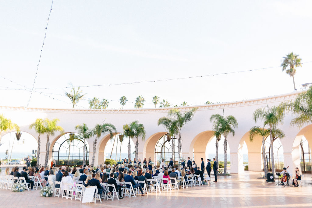 Outdoor event with many people seated under arches and palm trees, bright sky, and open space.