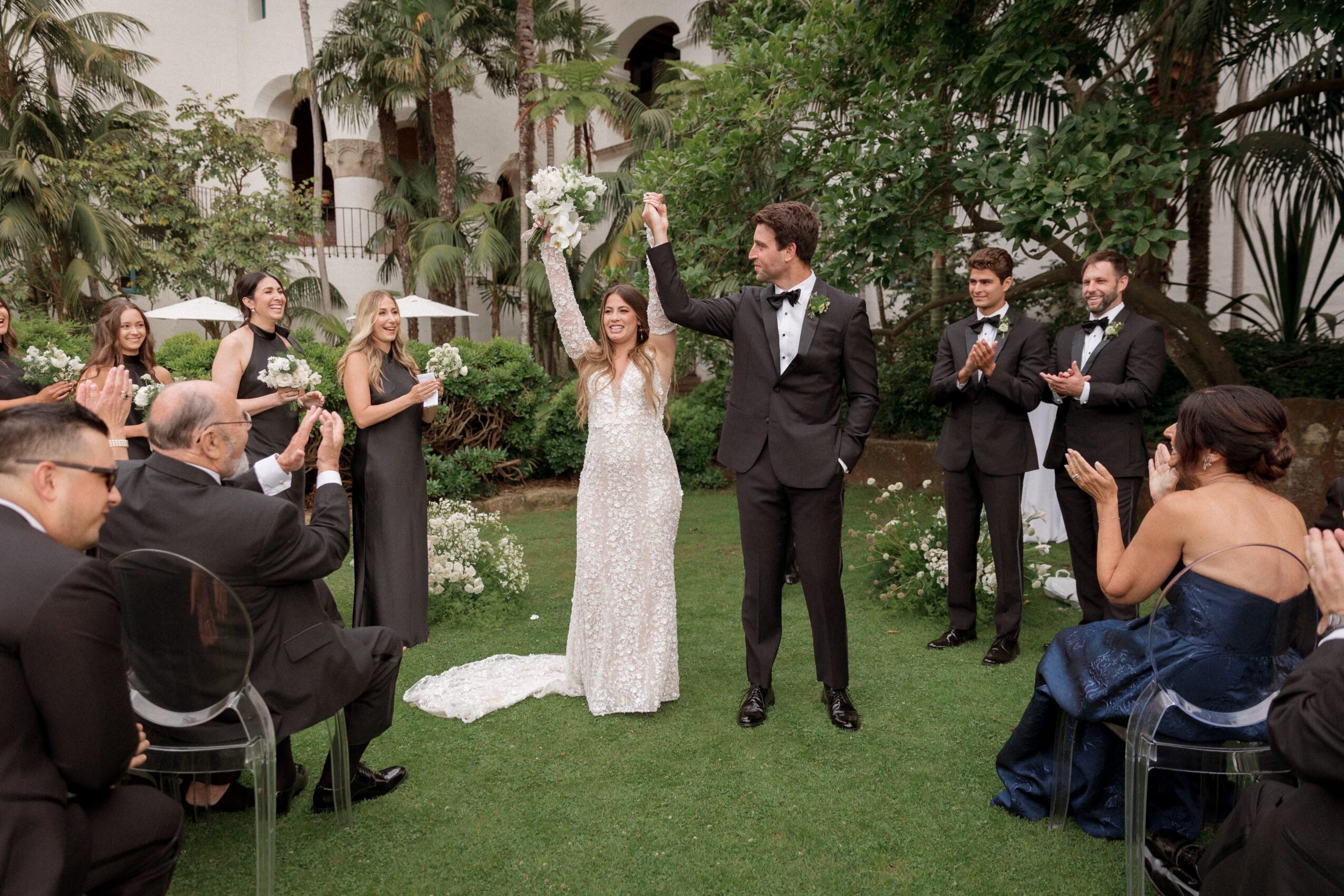 Bride and groom celebrating with guests outdoors, bride raising her hand, surrounded by trees and greenery.