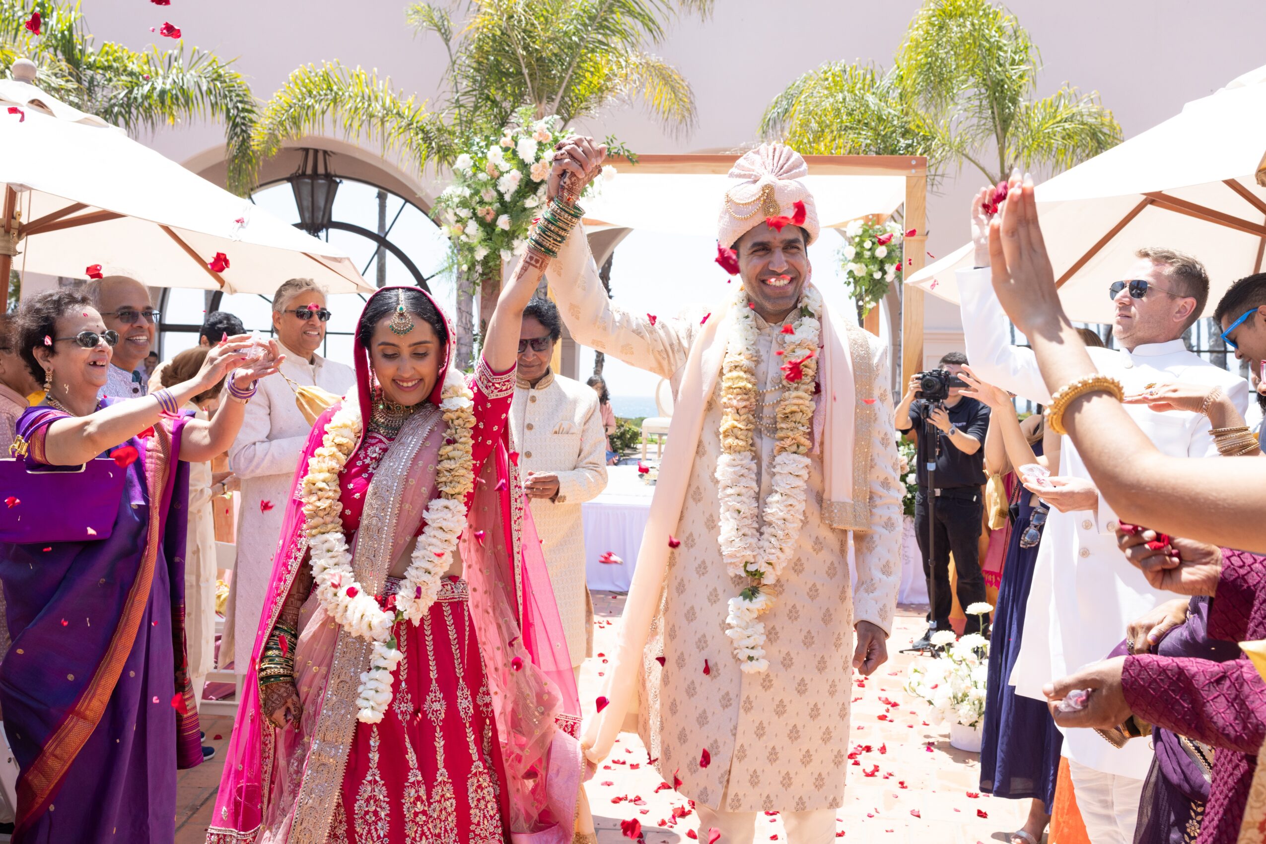 People celebrating a wedding outdoors with flower garlands, traditional attire, and joyful expressions, surrounded by umbrellas and greenery.