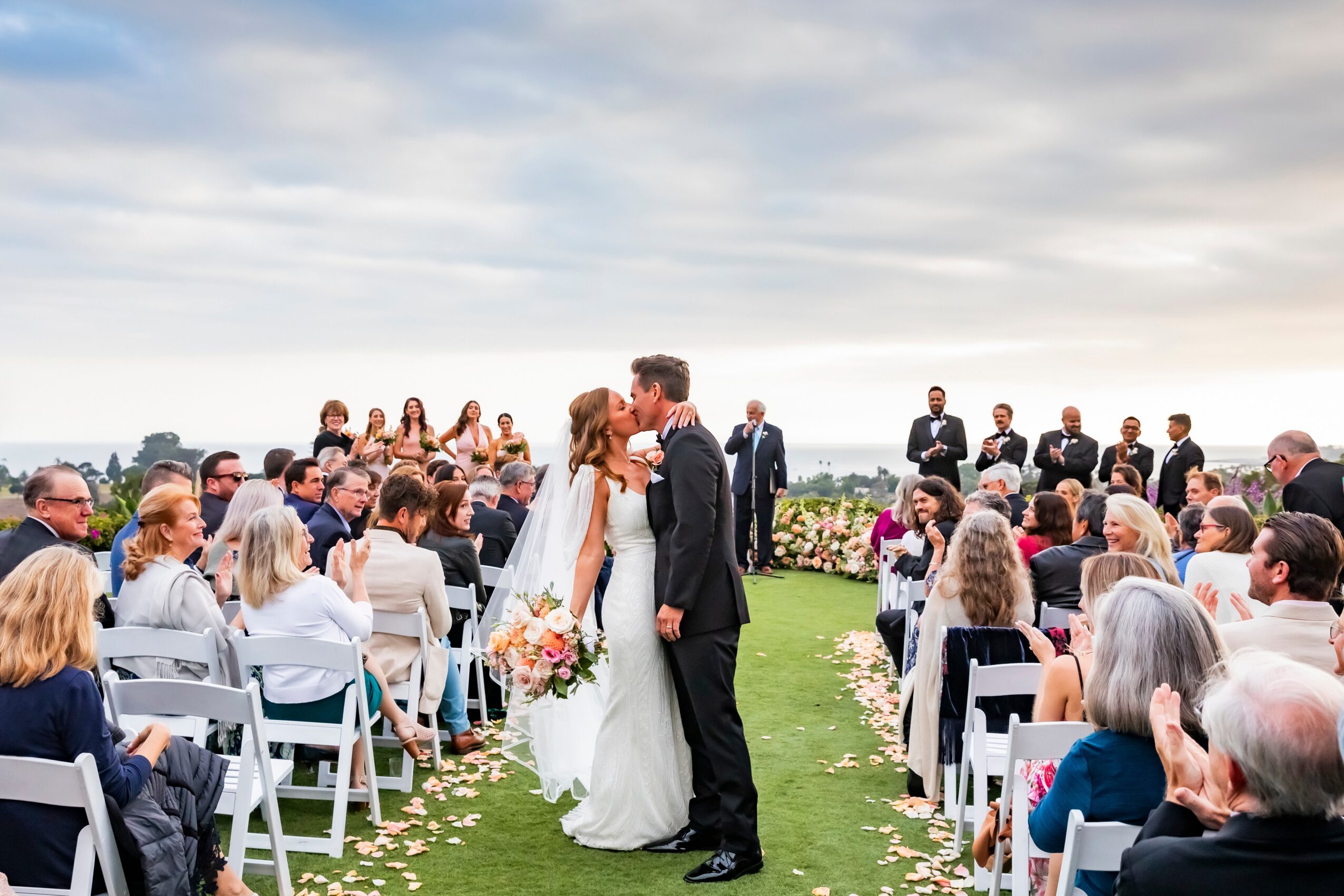 Bride and groom kiss during outdoor wedding ceremony with seated guests and a cloudy sky.