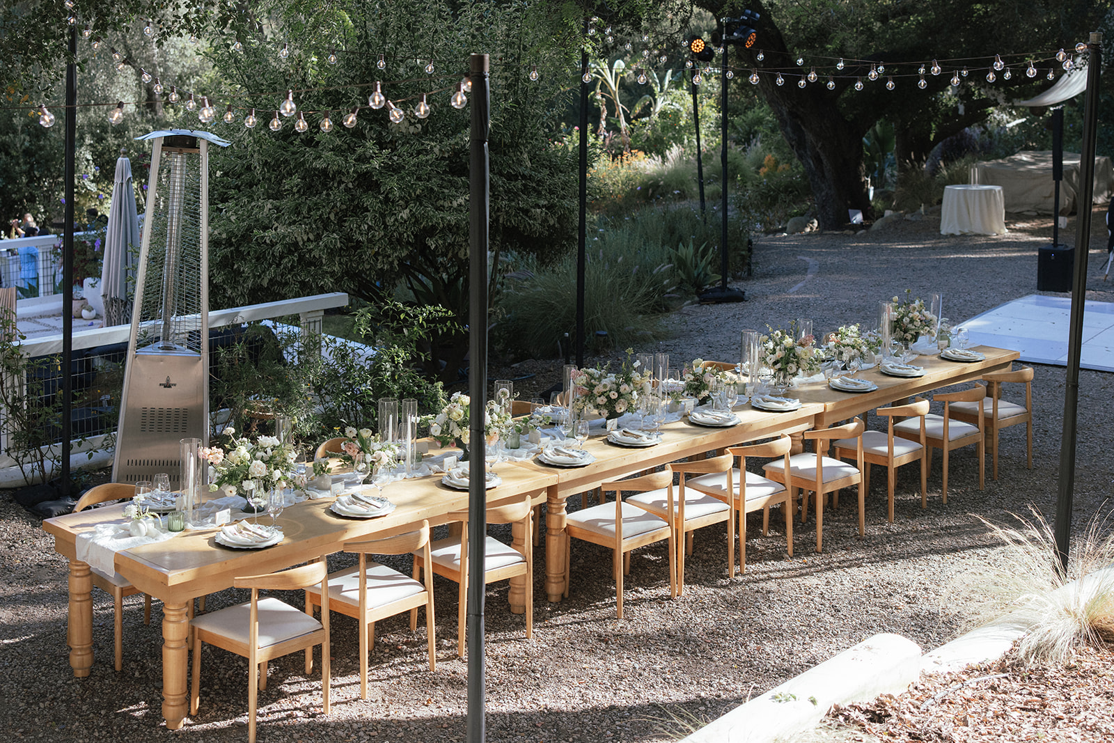 Long outdoor table set with plates, glasses, and floral centerpieces, surrounded by chairs, under string lights in a garden setting.