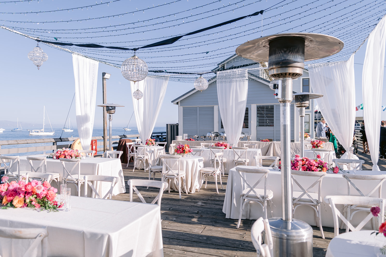 Outdoor dining area with white tables and chairs, decorated with pink flowers, under a clear sky near water.