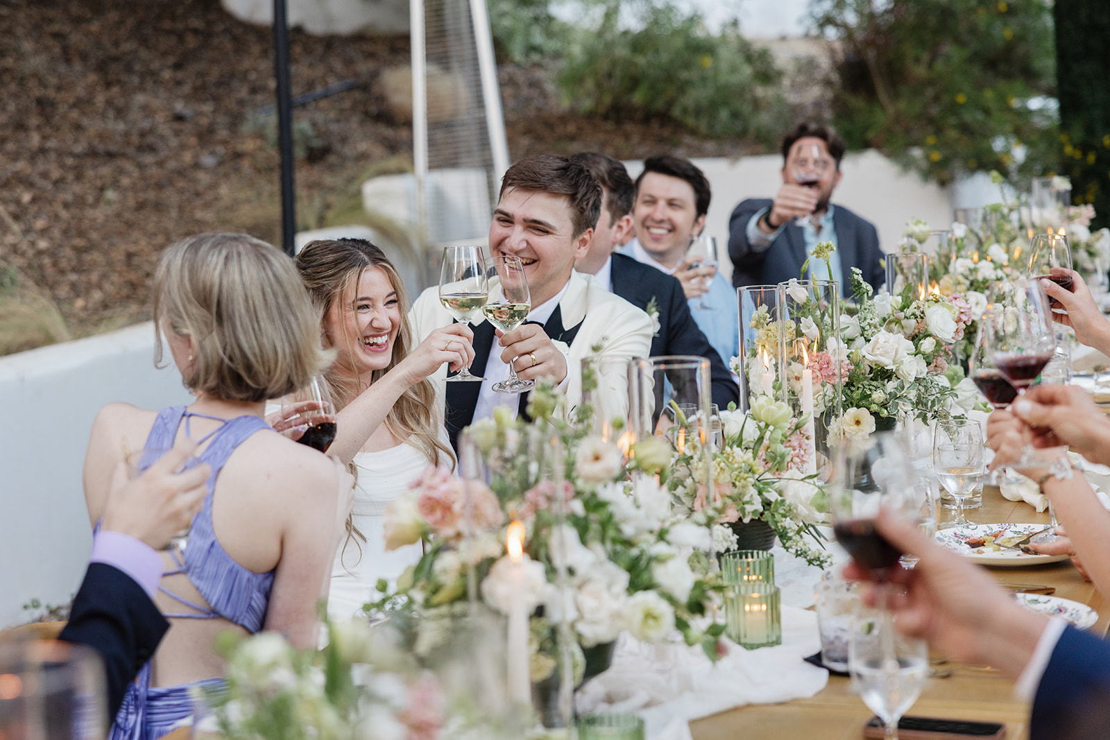 People celebrating at a table with flowers, drinks, and smiling faces outdoors.