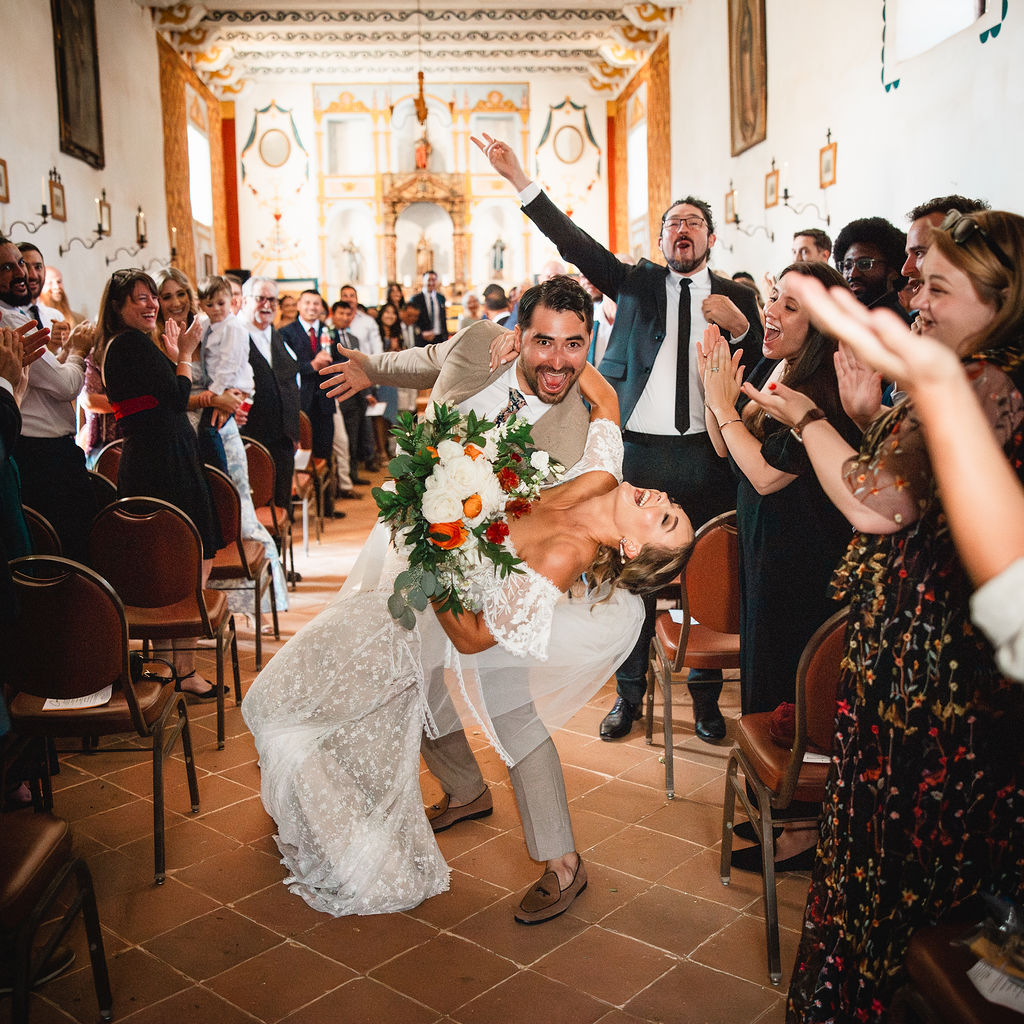Couple dancing in a decorated church, surrounded by cheering guests, with ornate interior and high ceiling.