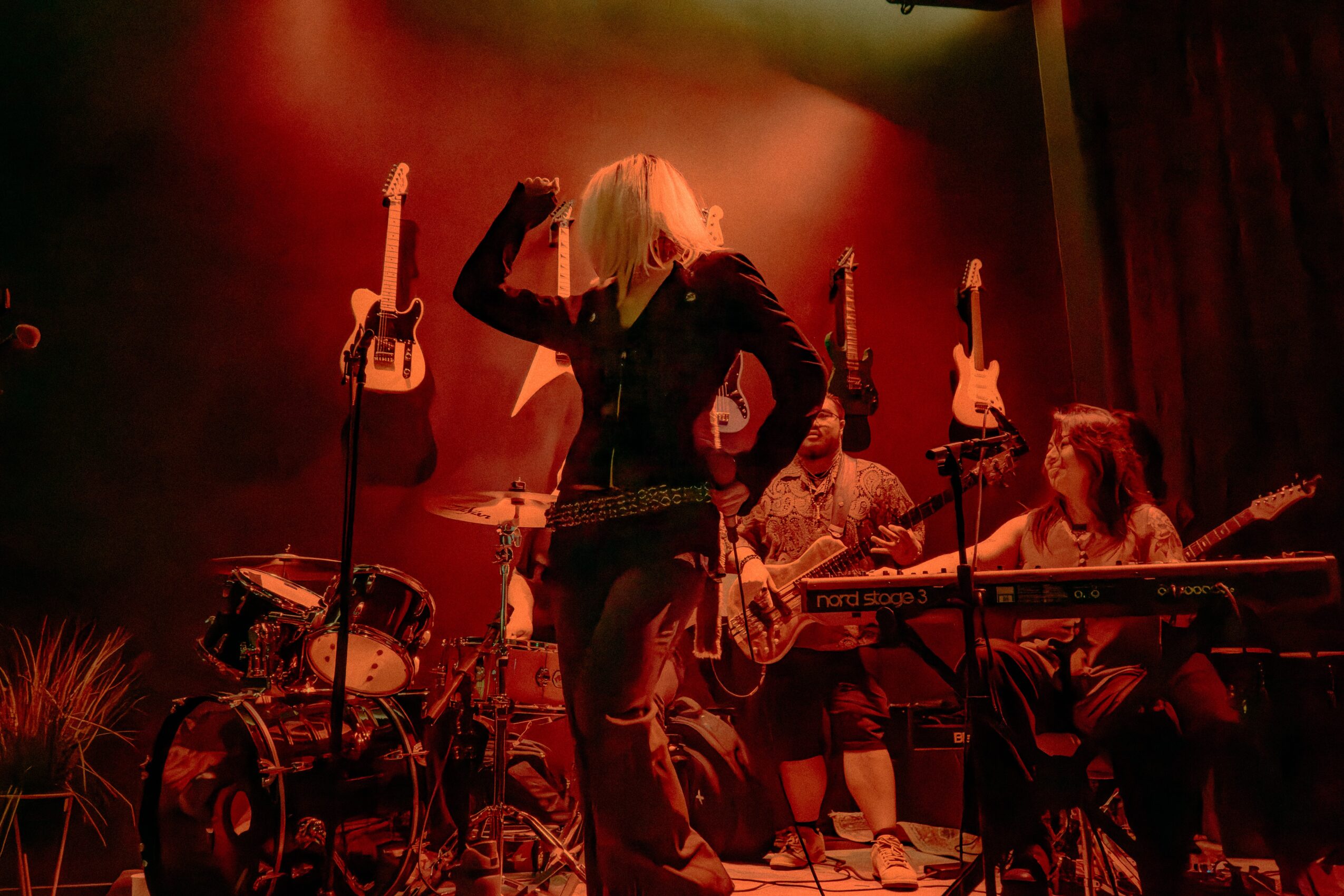 Musicians perform on stage with guitars, keyboard, and drums, illuminated by red and orange lighting, with a female singer in the center.