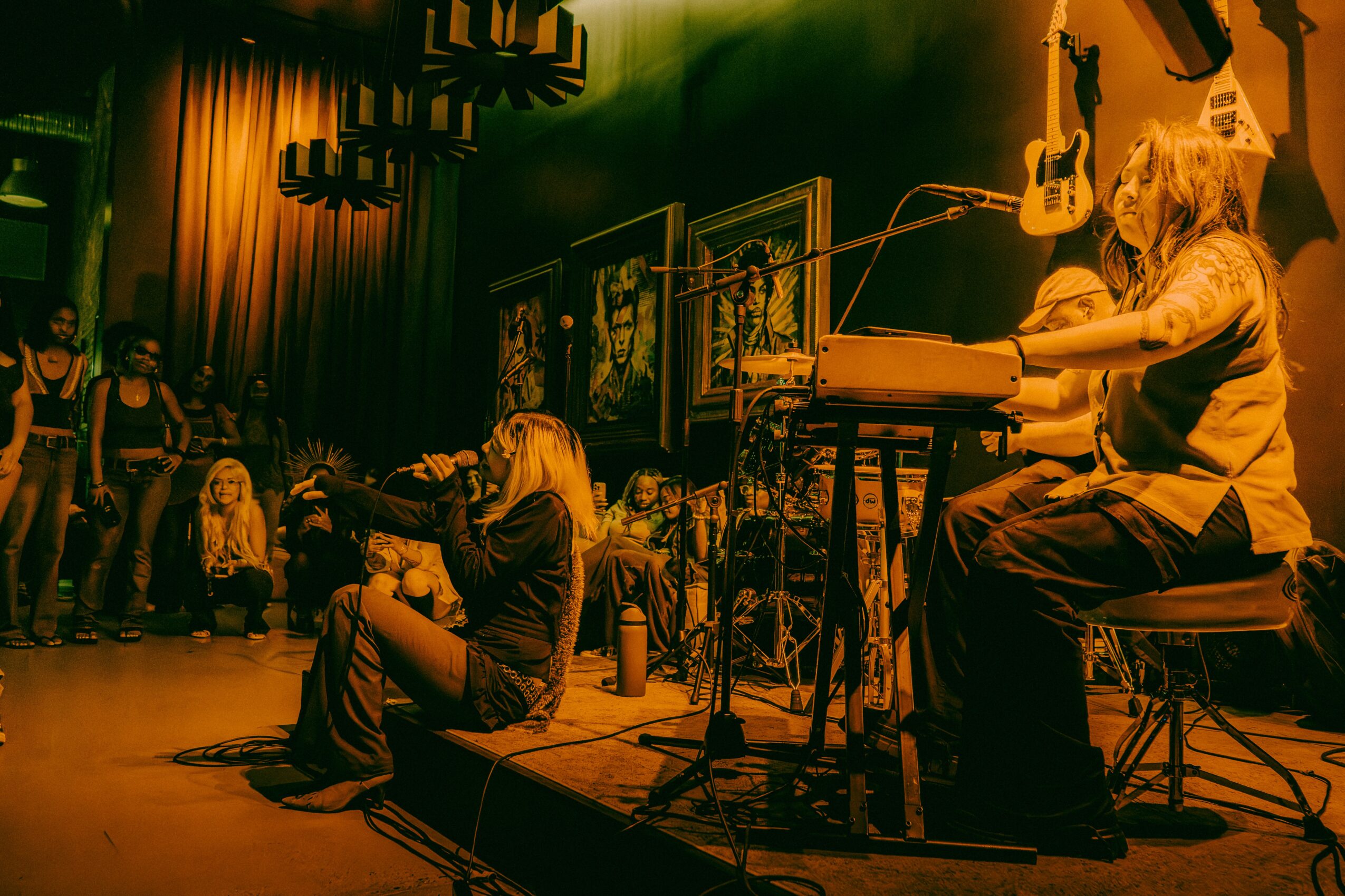 Musicians perform on stage with guitars and keyboard, audience watching in a dimly lit venue, yellow and green lighting.