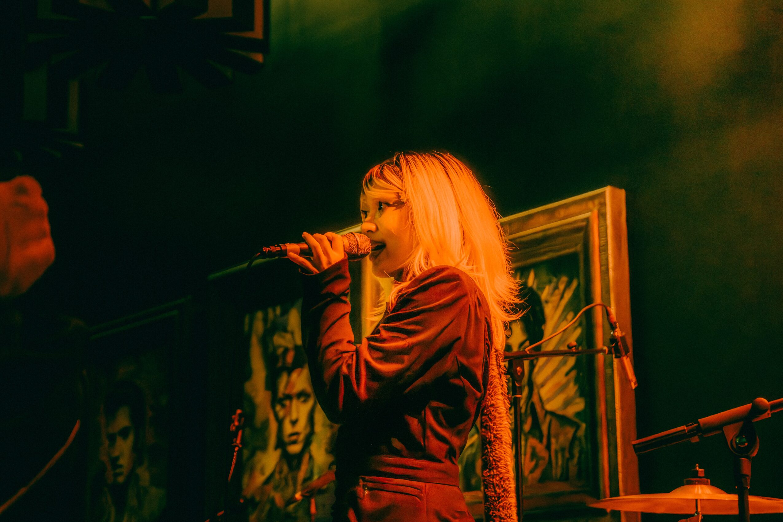Person with blonde hair singing into a microphone on stage, with framed artwork in the background, dim lighting.