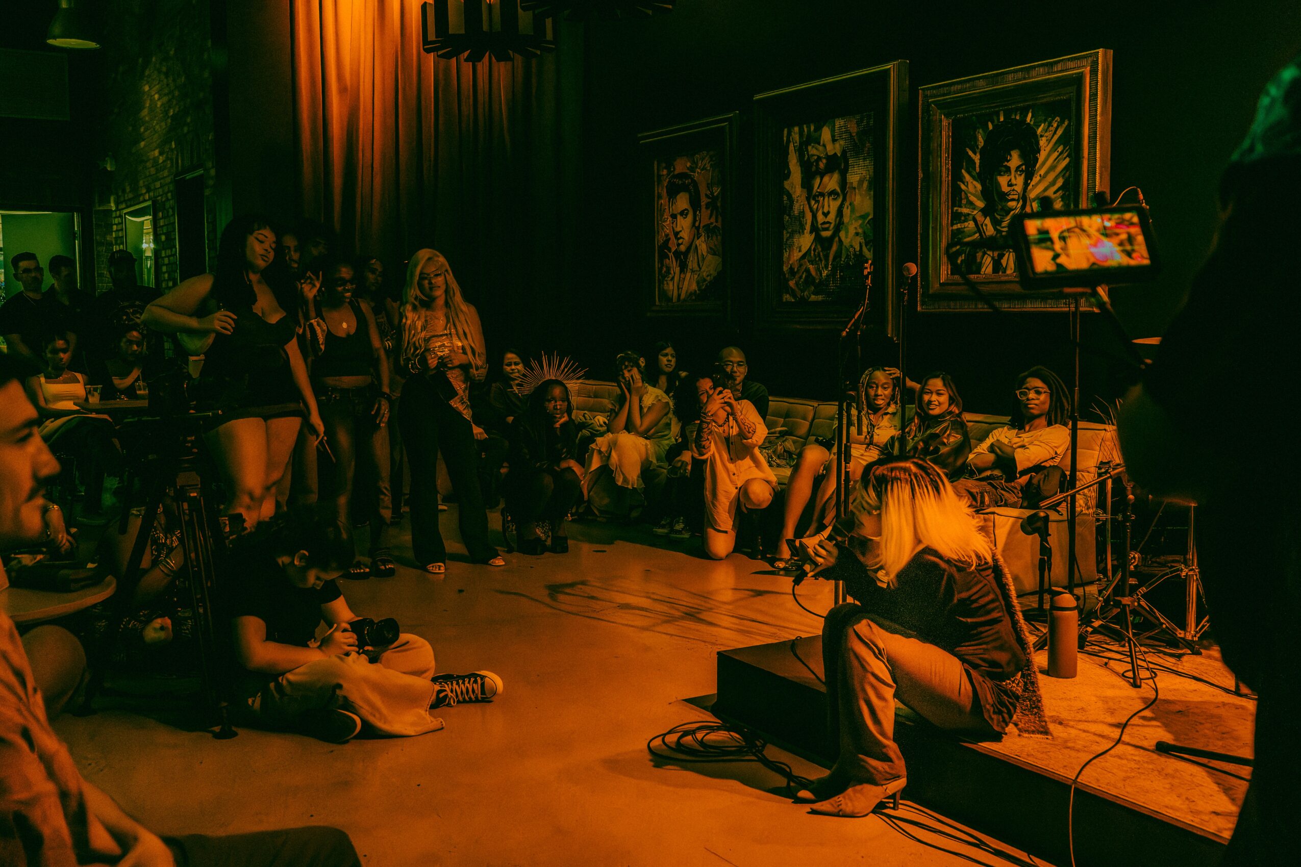 People gathered in a dimly lit room with framed artwork on the wall, some sitting and others standing, with a musician playing guitar.