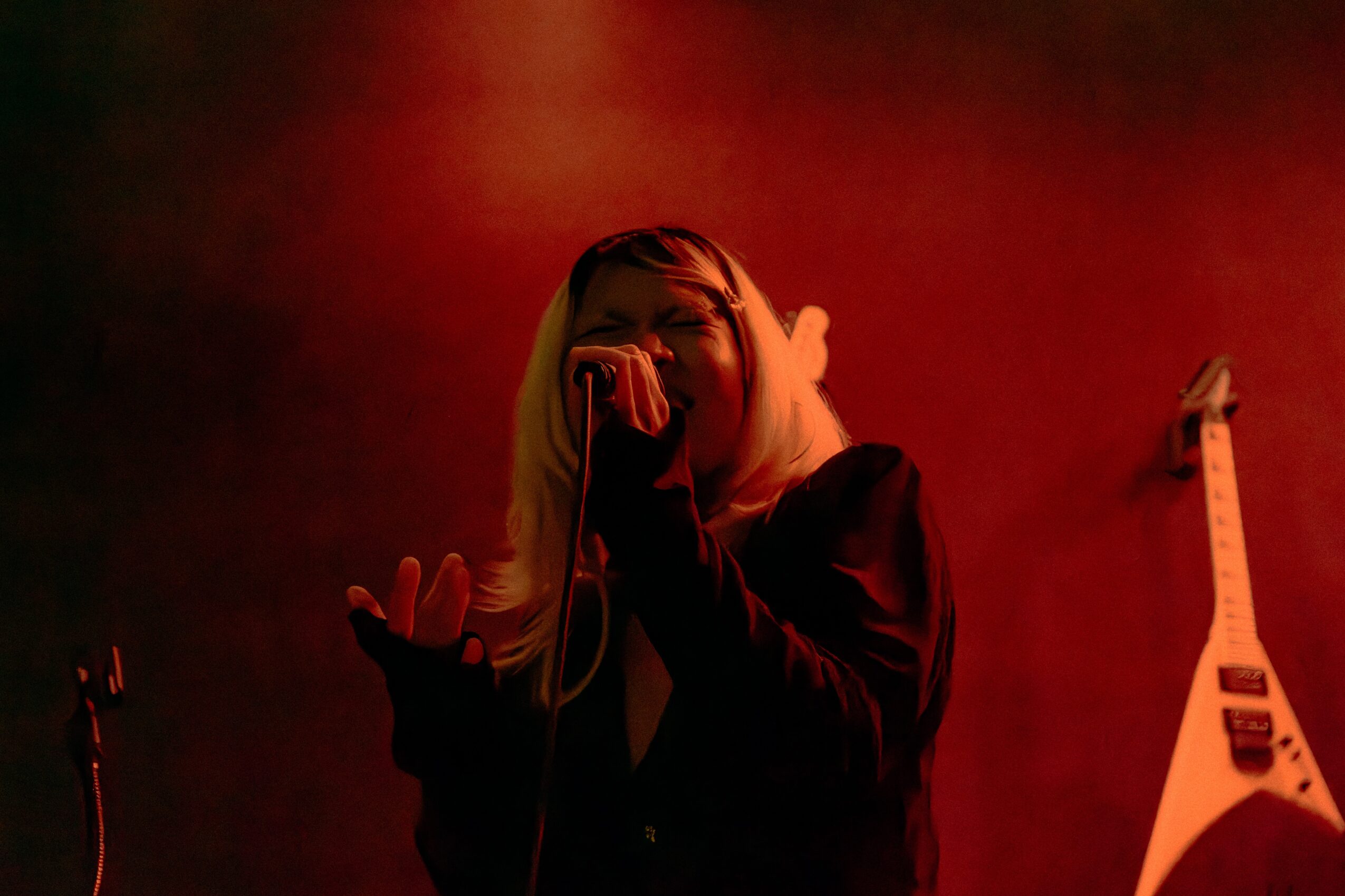 Person with long hair holding a microphone, performing on stage with red lighting, guitar on the wall behind.