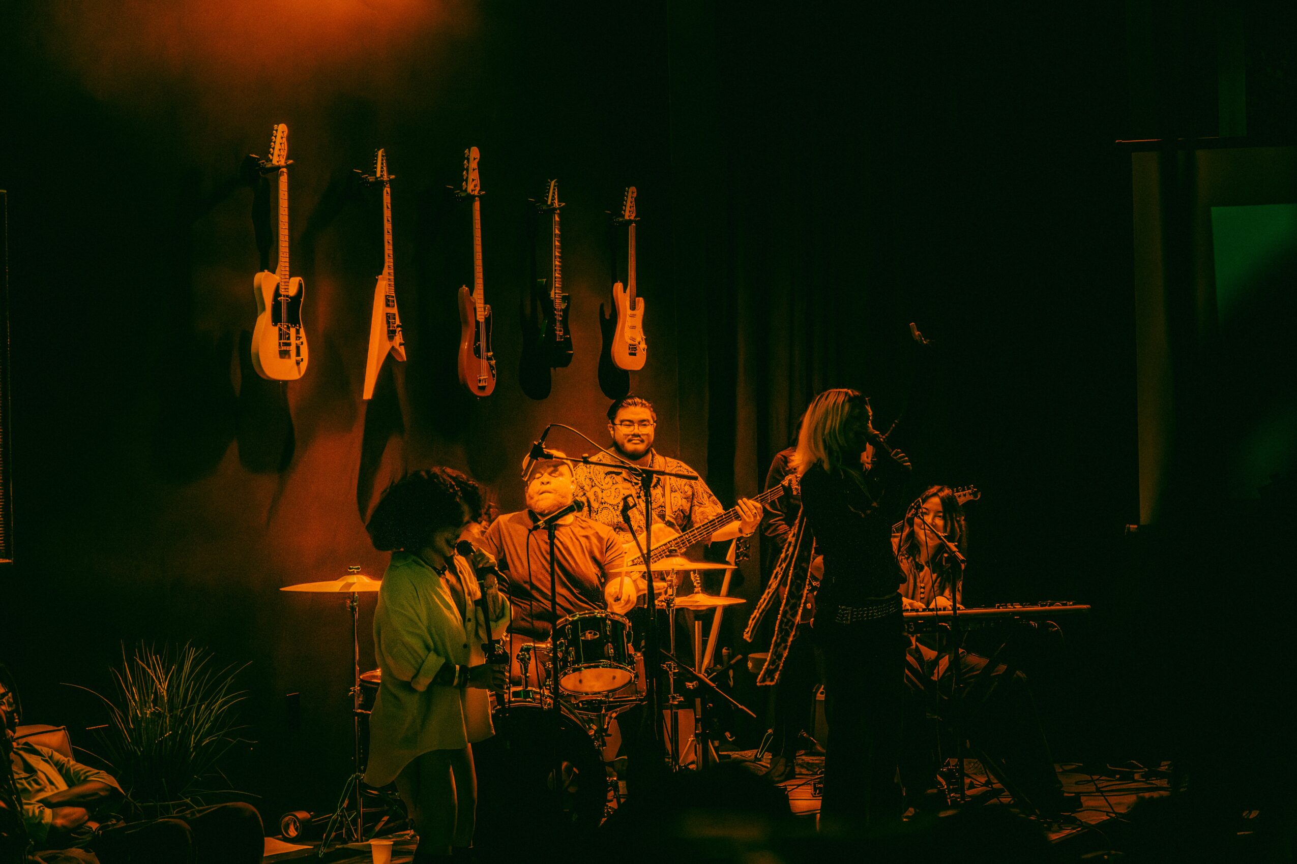 Musicians perform on stage with guitars hanging in the background, dim lighting, and a person playing drums.
