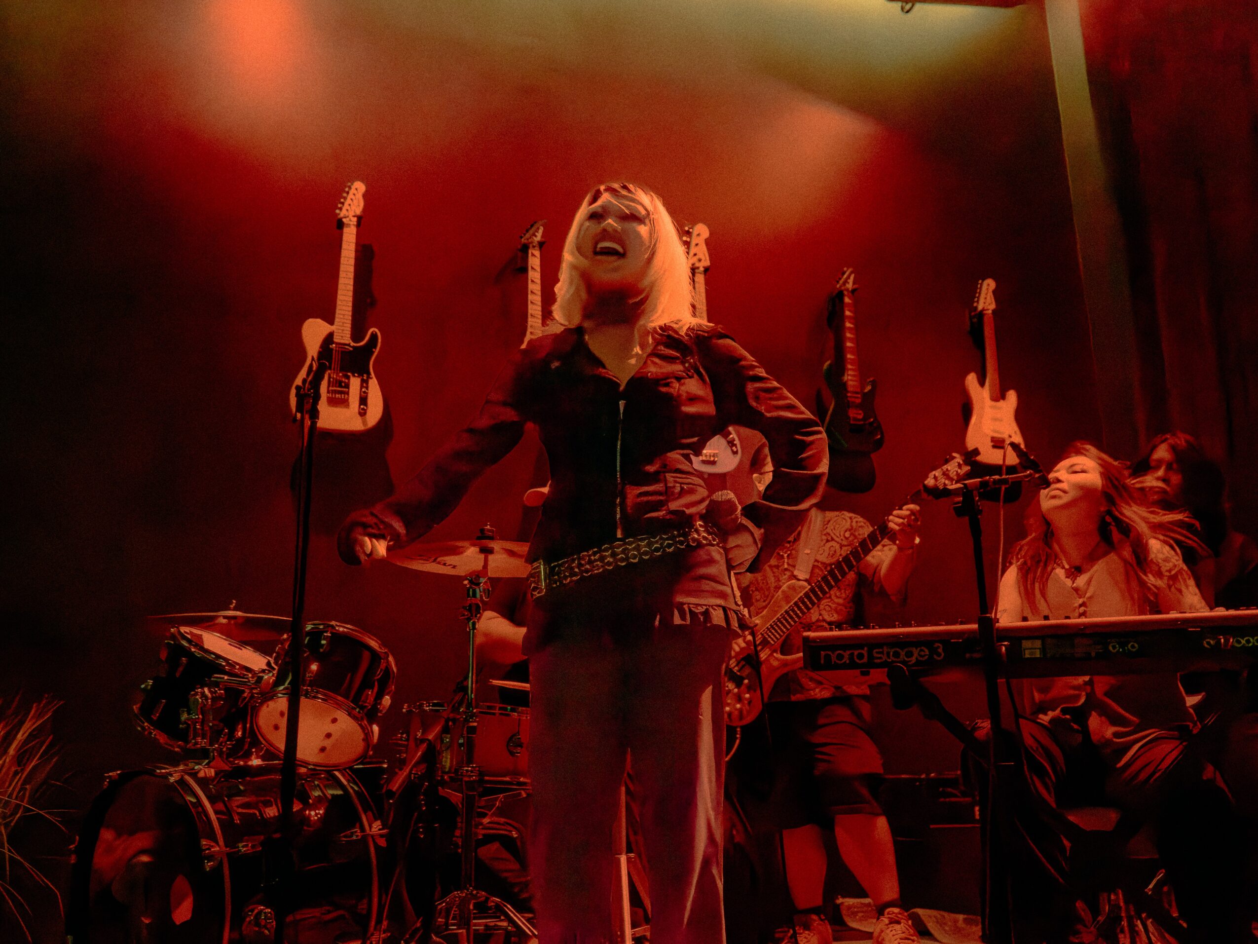 A woman on stage with musical instruments, smiling and raising her arms, with a red background.