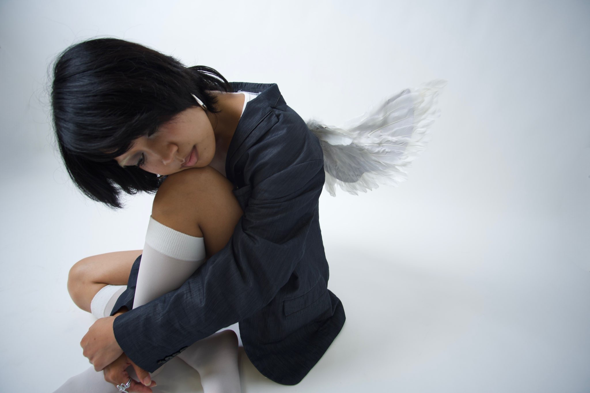 Person with black hair sitting with knees drawn up, wearing a dark jacket and white thigh-high socks, against a plain background.