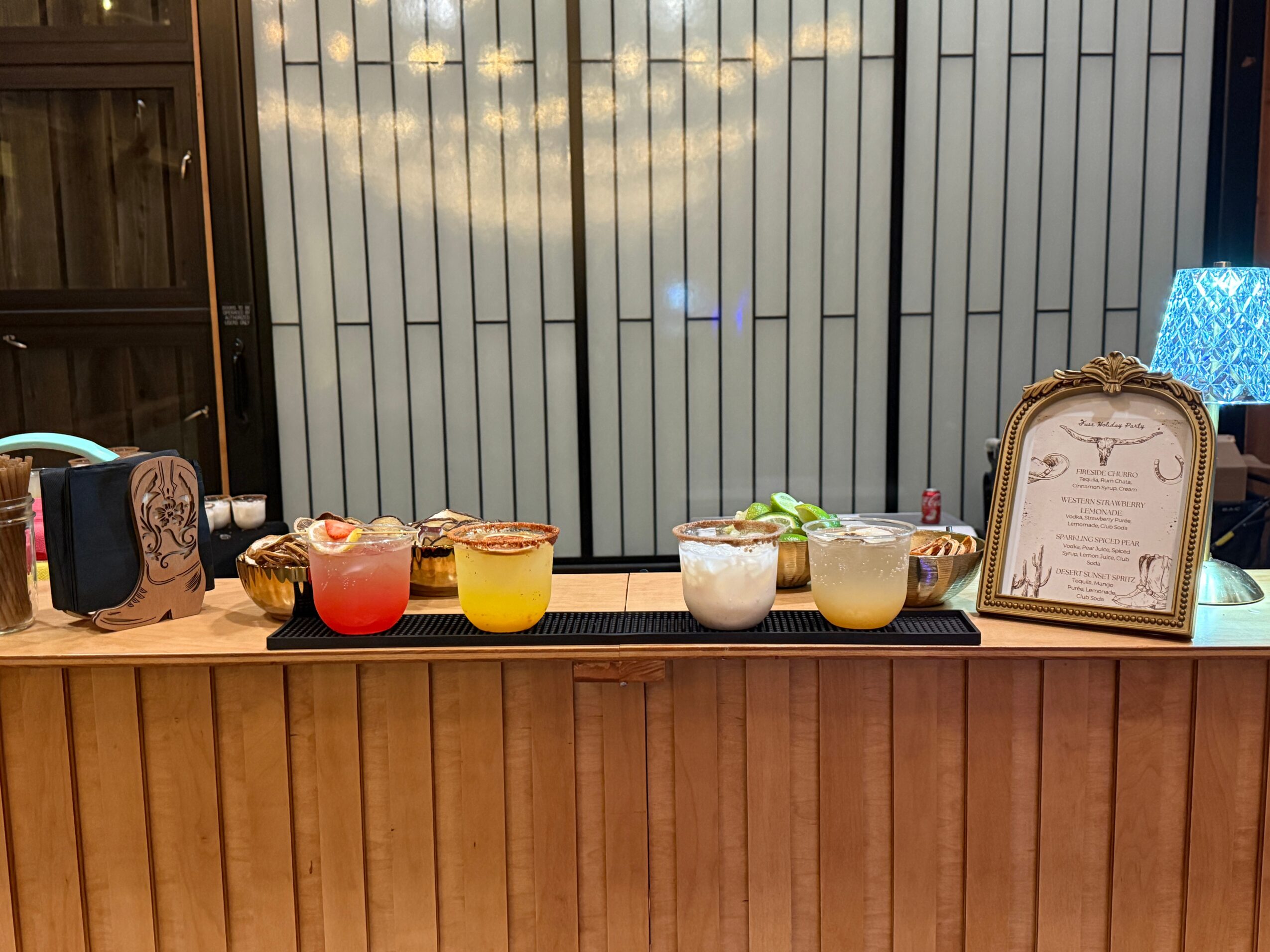 Bar counter with colorful drinks, framed menu, and decorative lamp, set against a window with vertical blinds.