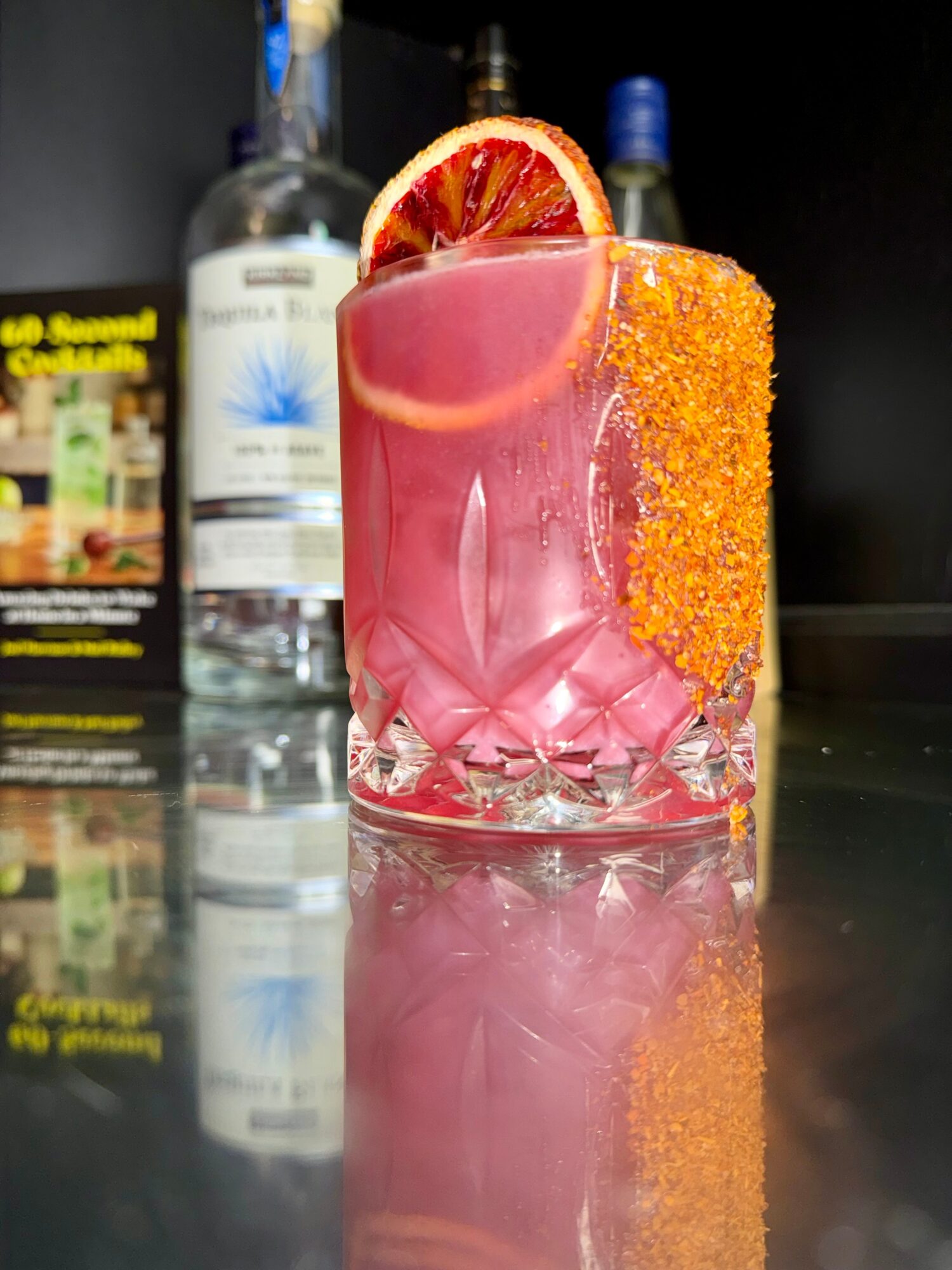 Pink cocktail with orange sugar rim and blood orange slice, ice cubes visible, on a reflective surface.