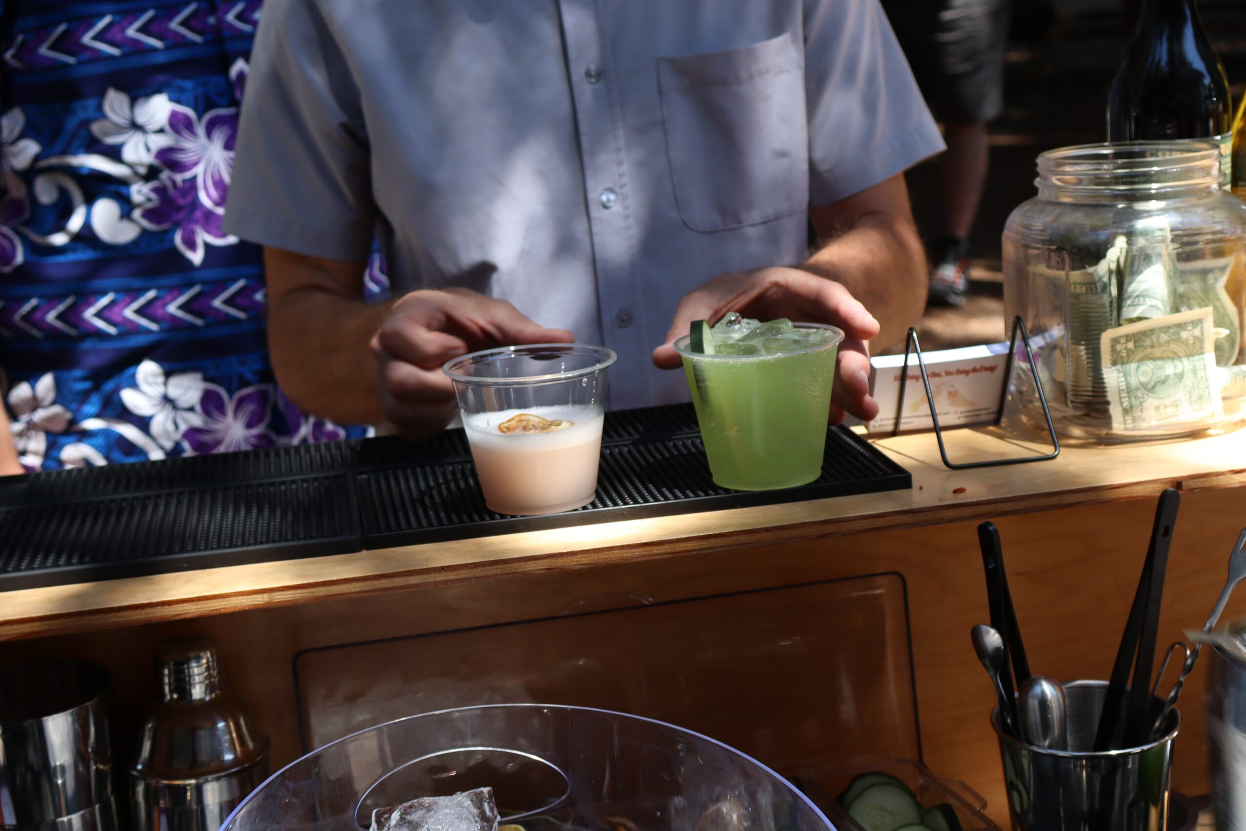 Person standing behind a bar with drinks and jars, with a person in a floral shirt partially visible on the left.