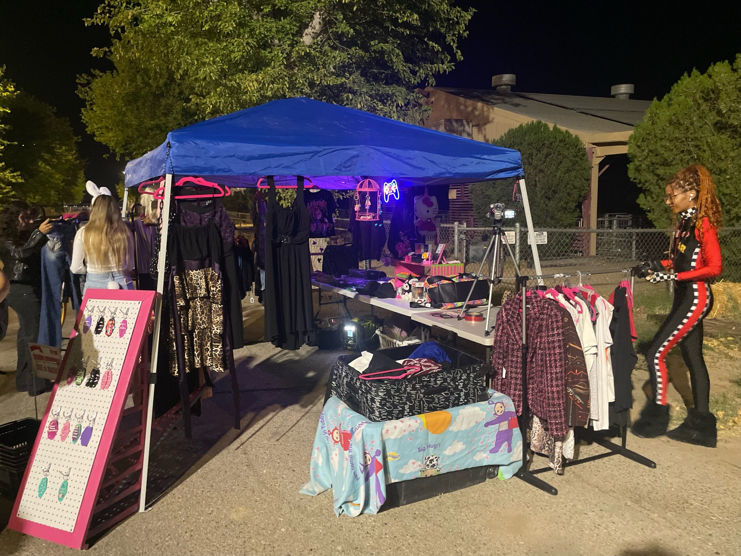 Nighttime outdoor market stall with clothing under a blue canopy, people browsing, and a woman in red and black outfit nearby.